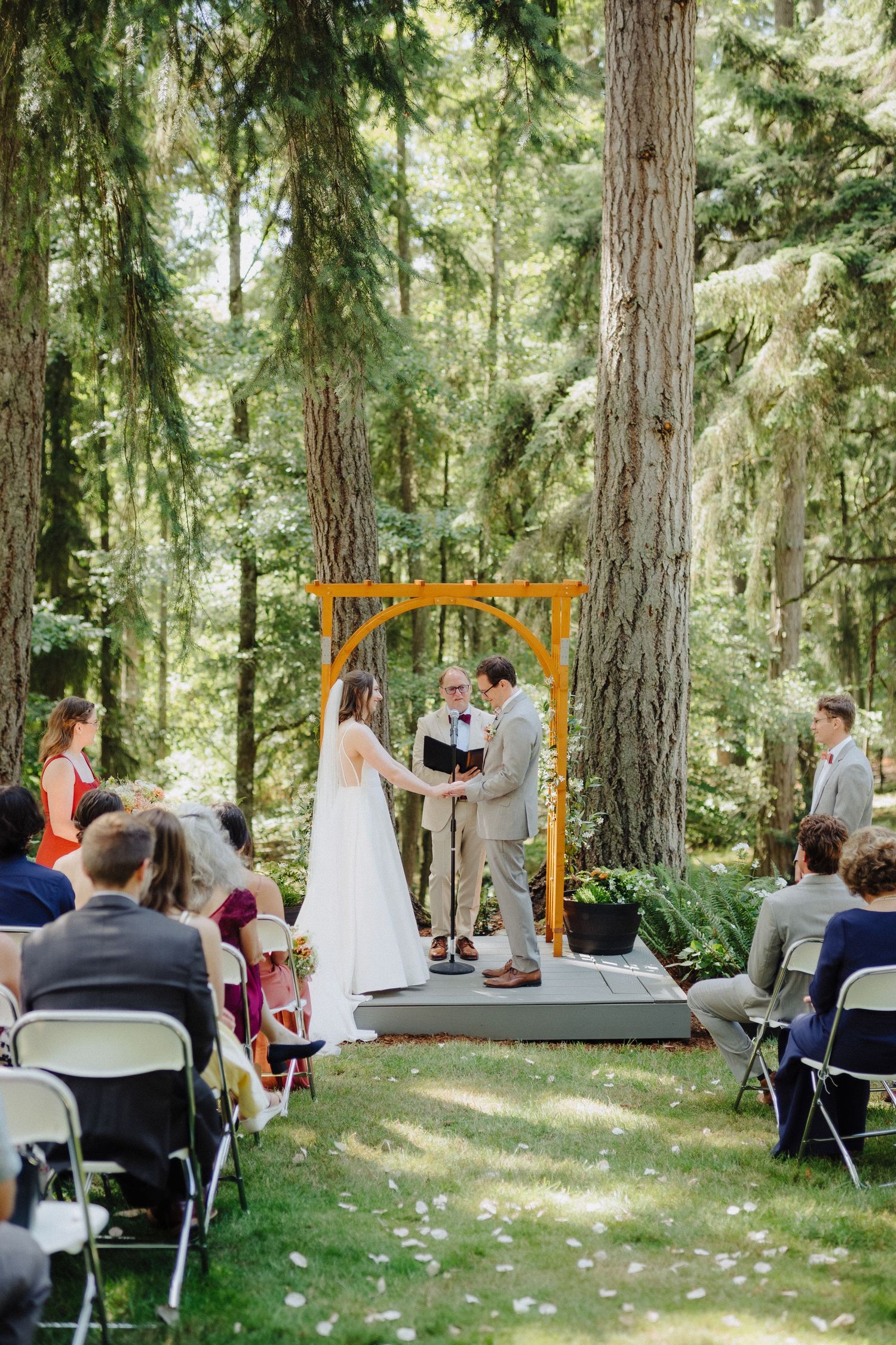 Outdoor forest wedding ceremony with guests seated on white chairs under tall evergreen trees.