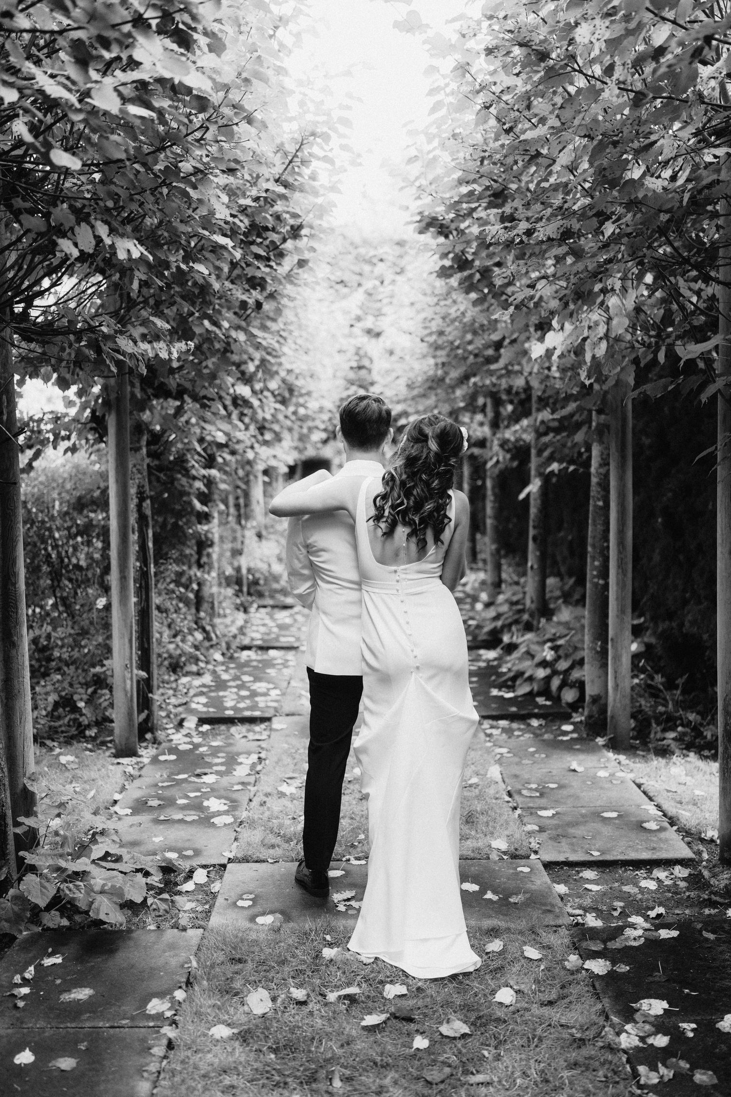 A couple in wedding attire stands in a romantic garden pathway lined with trees and fallen leaves.