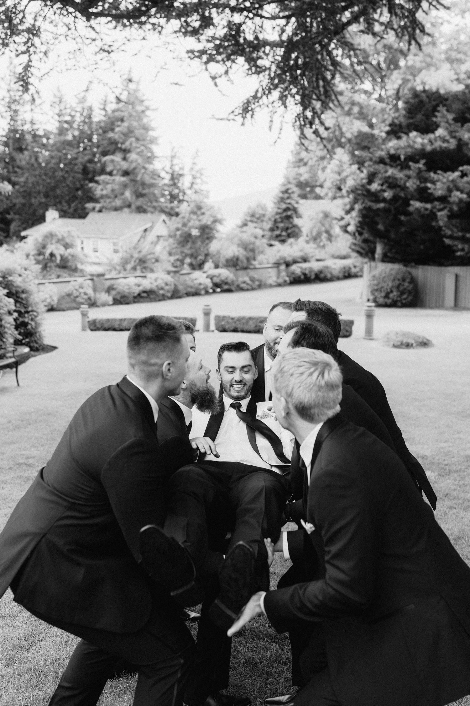 Groomsmen in black suits huddle together playfully on a grassy lawn.