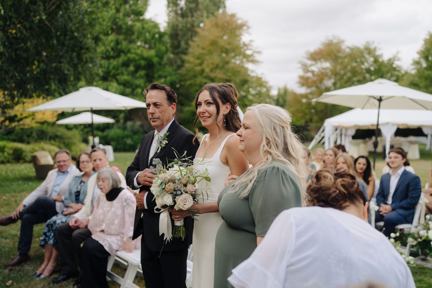 Wedding party celebrates outdoors under white tents and umbrellas in a garden setting.