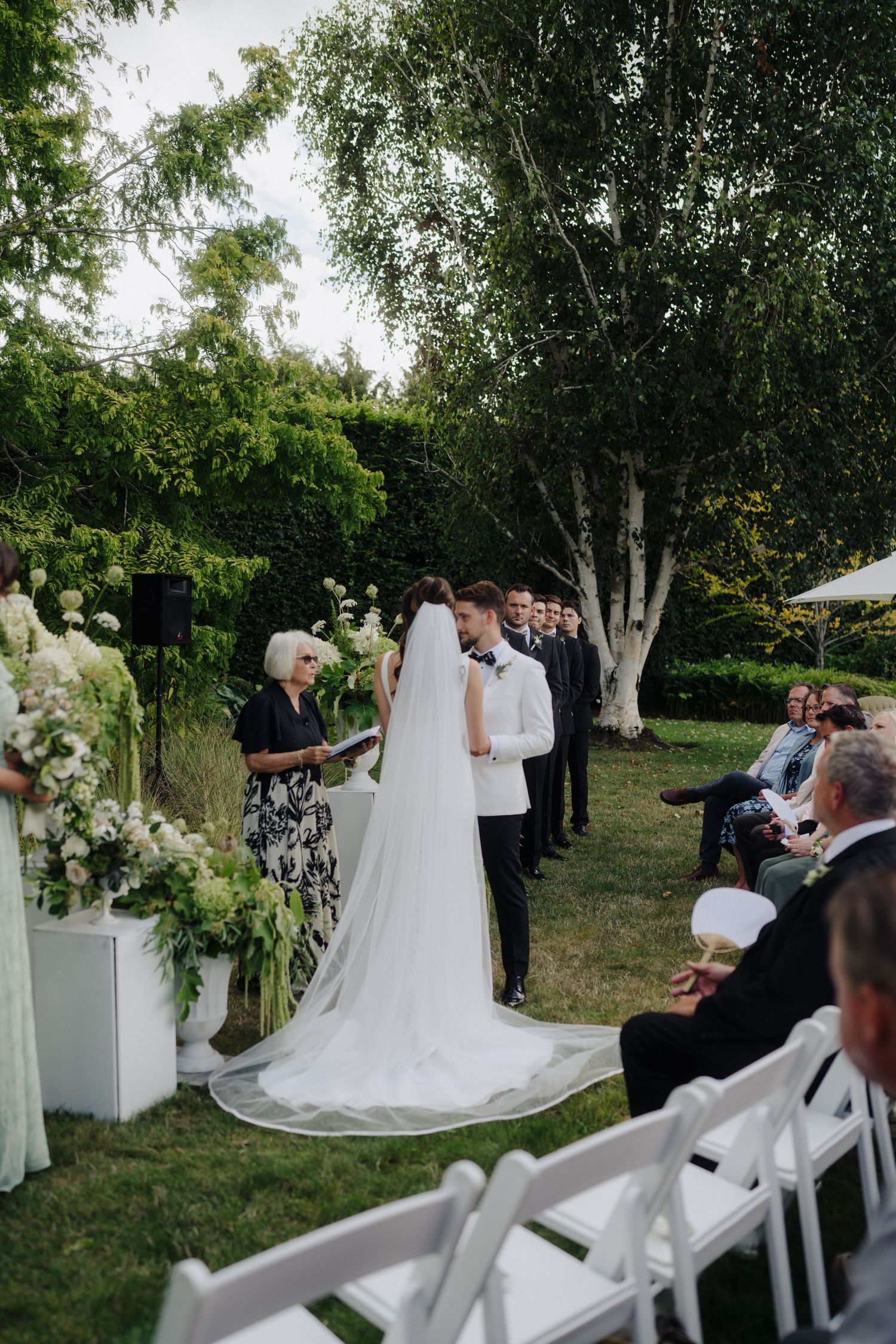 Outdoor wedding ceremony with white chairs and elegant floral decorations in a garden setting.