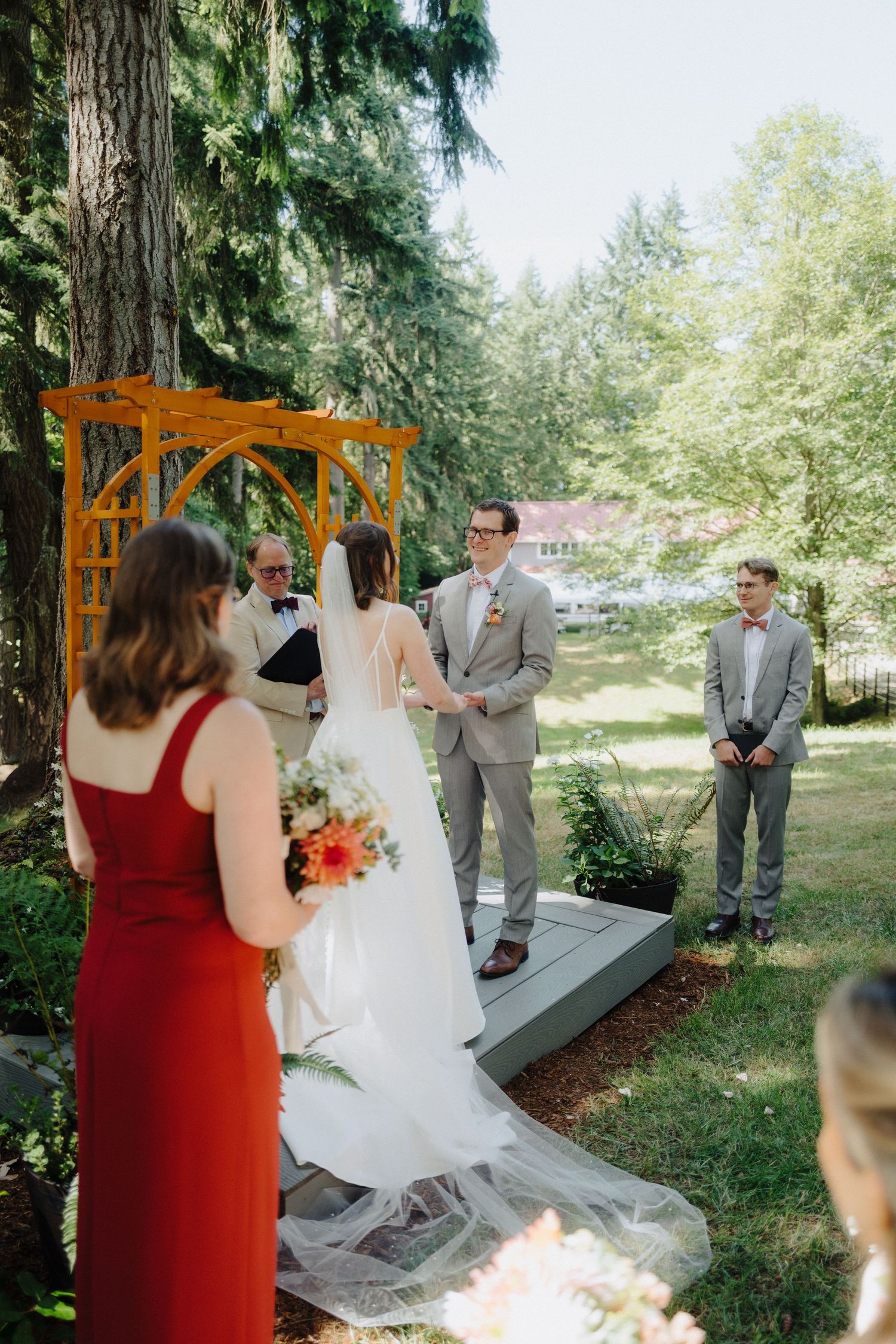 An outdoor wedding ceremony takes place under tall evergreen trees with guests seated on wooden benches.