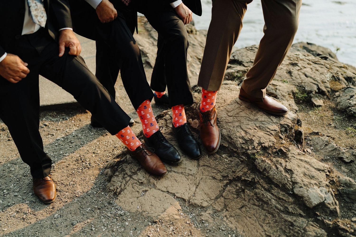 Groomsmen showing off their matching red patterned socks while standing on rocky coastal terrain.
