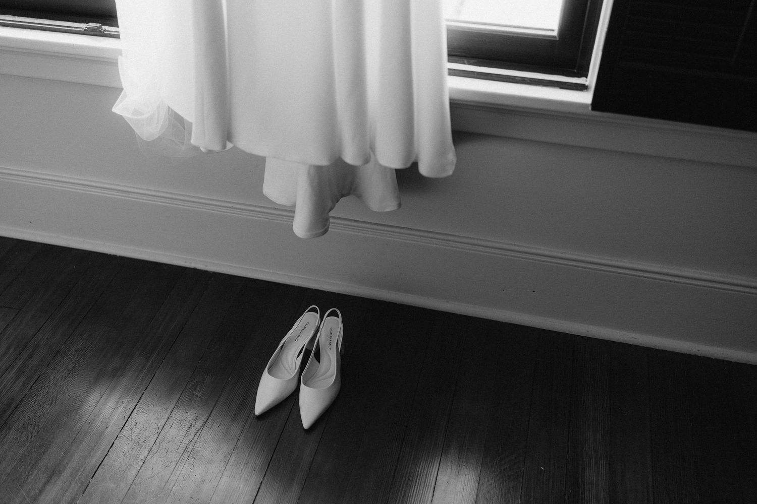 A white wedding dress hangs by a window with ballet shoes placed on dark hardwood floor below in black and white.