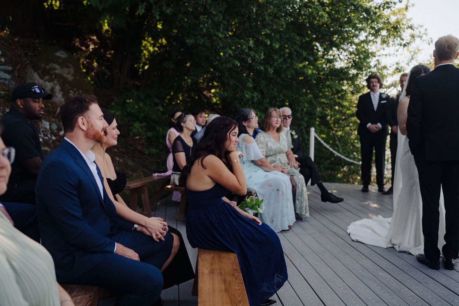 Wedding guests seated on wooden deck during outdoor ceremony with natural greenery backdrop.