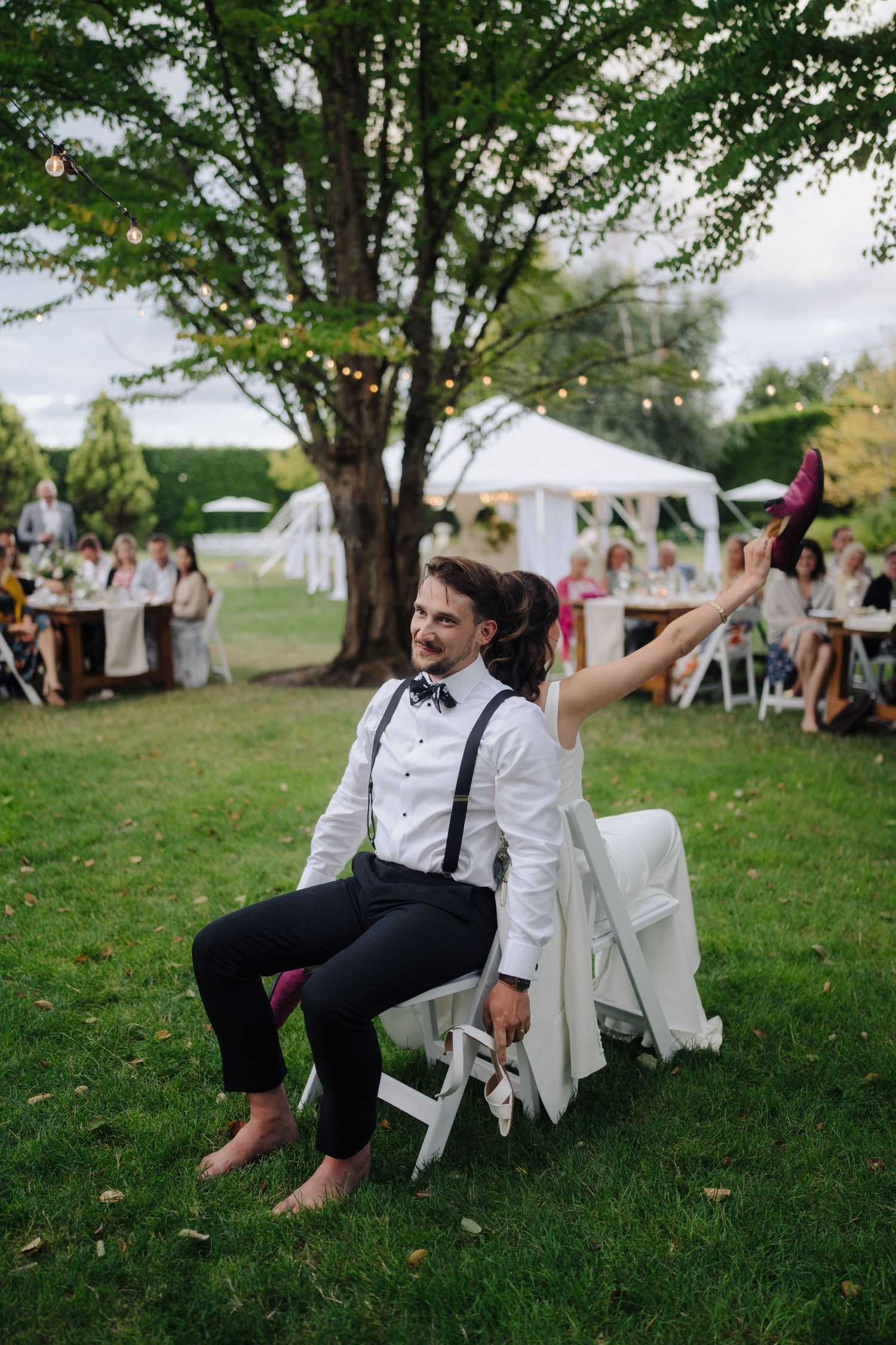 Wedding couple relaxes in white chairs on grassy lawn during outdoor reception.