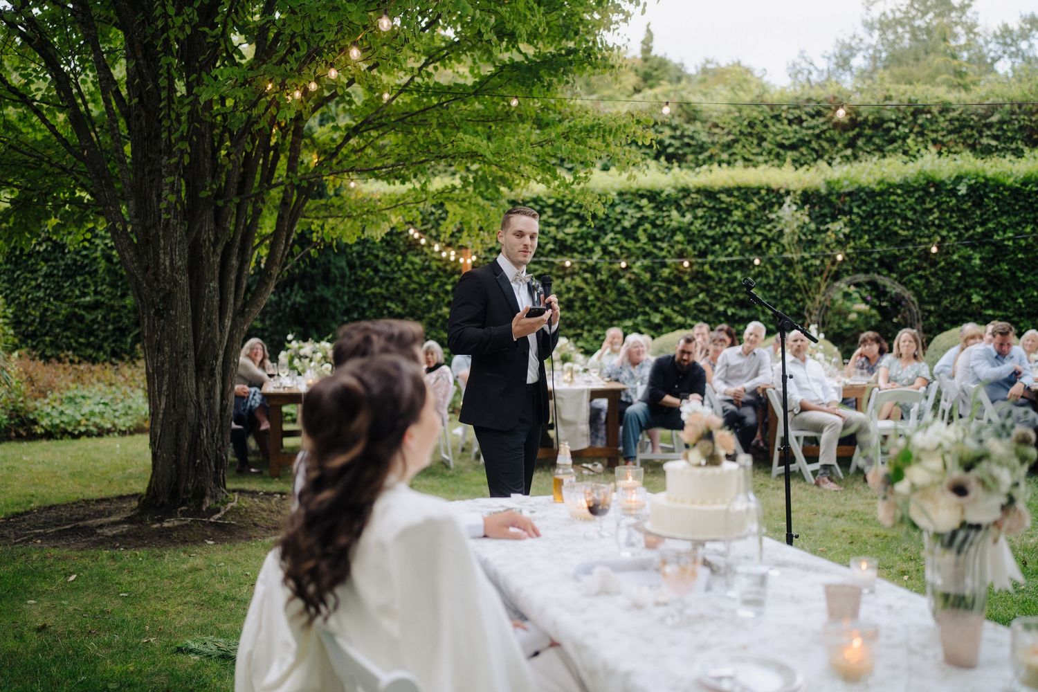 Garden wedding reception with white-clothed tables arranged under green trees.