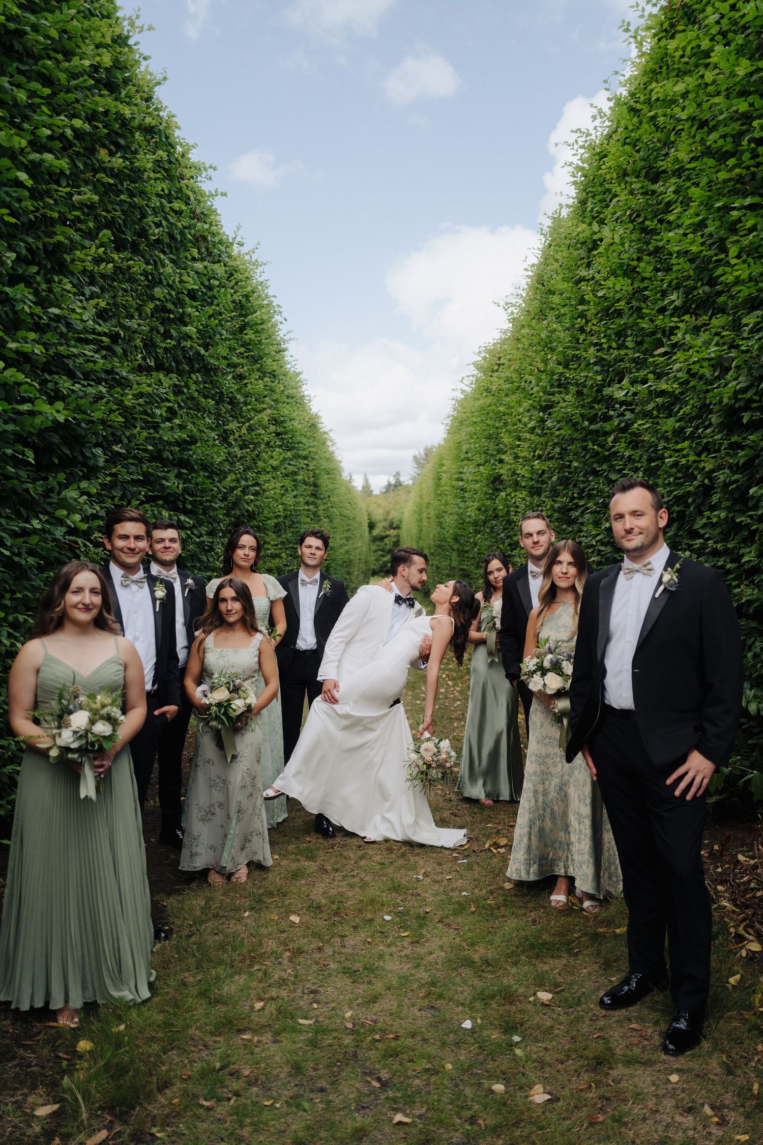 Wedding party poses in a beautiful garden pathway lined with tall green hedges on a sunny day.