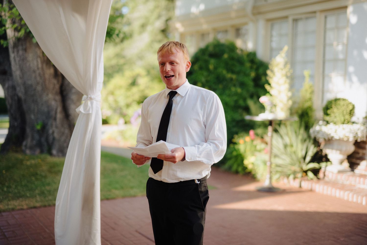 Person in white shirt and tie reads from paper during outdoor ceremony.