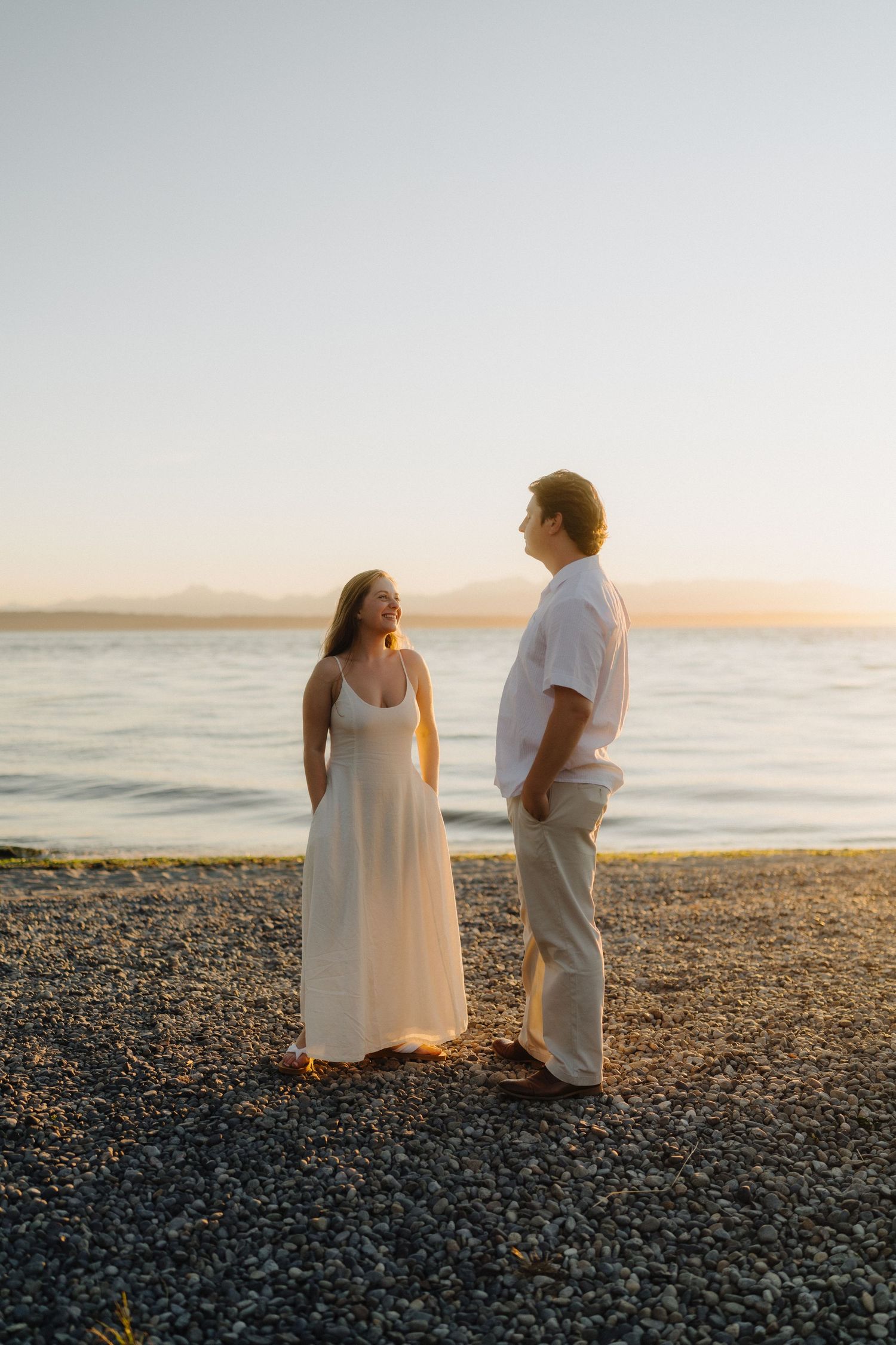 Two figures in white clothing stand together on a rocky beach at sunset against a peaceful ocean backdrop.