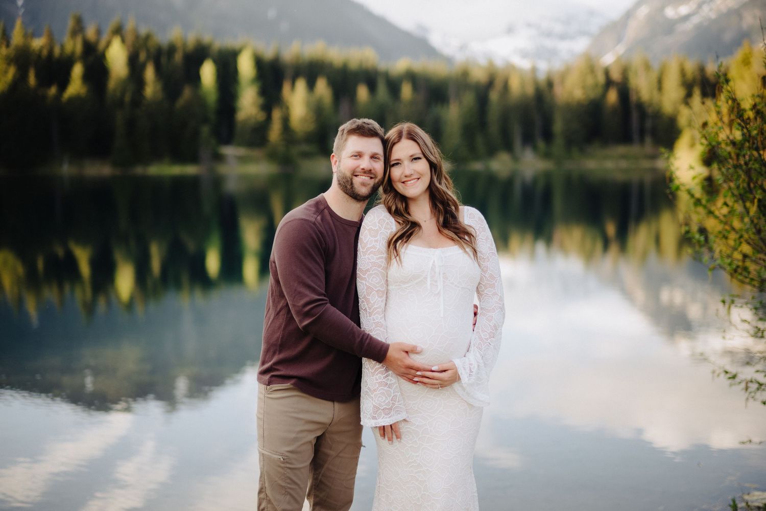 Mountain lake landscape with evergreen trees reflects in calm waters as couple shares intimate moment at sunset.