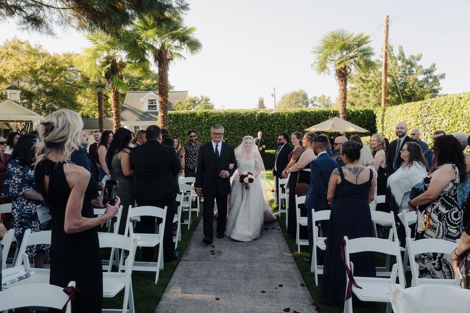 Wedding ceremony procession down an aisle lined with white chairs and guests in formal attire.
