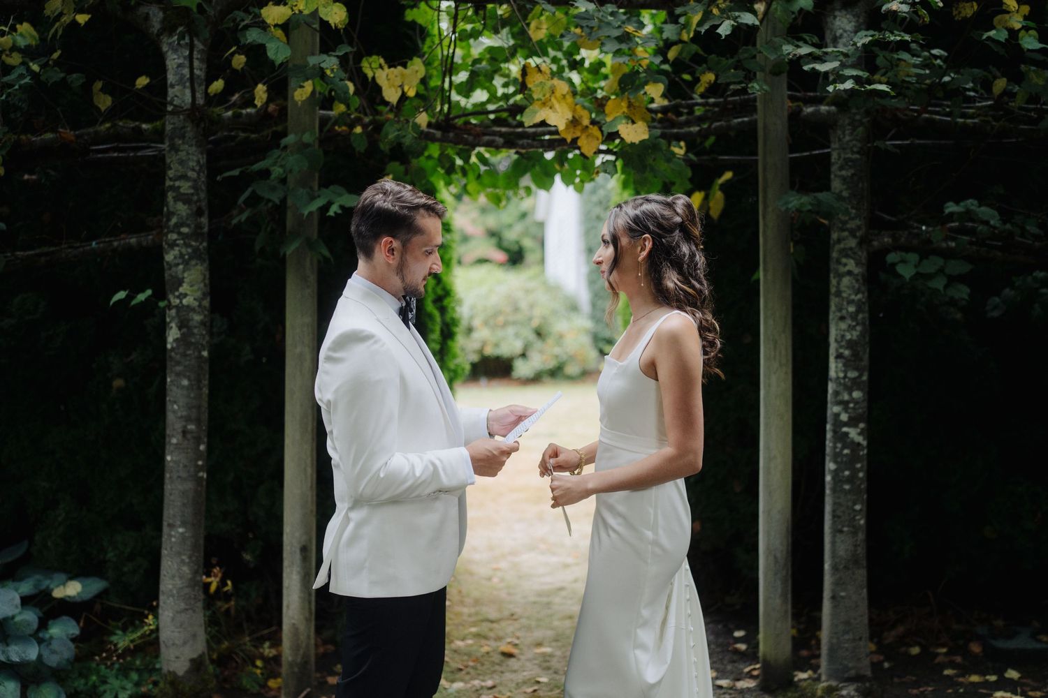 Wedding couple holding hands during outdoor ceremony beneath natural archway of trees.