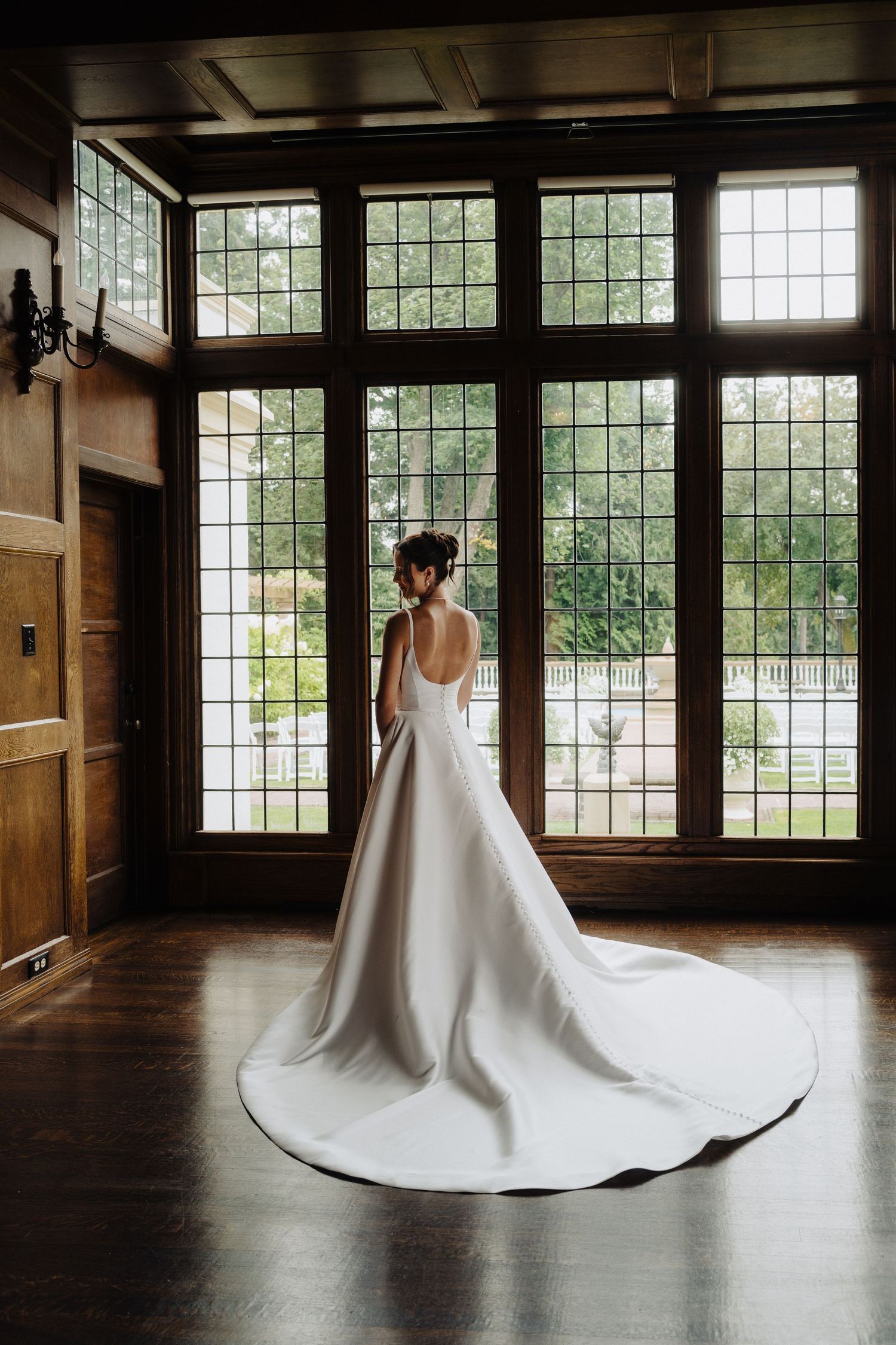 An elegant white wedding dress with a dramatic train displayed in a wood-paneled room with large windows.