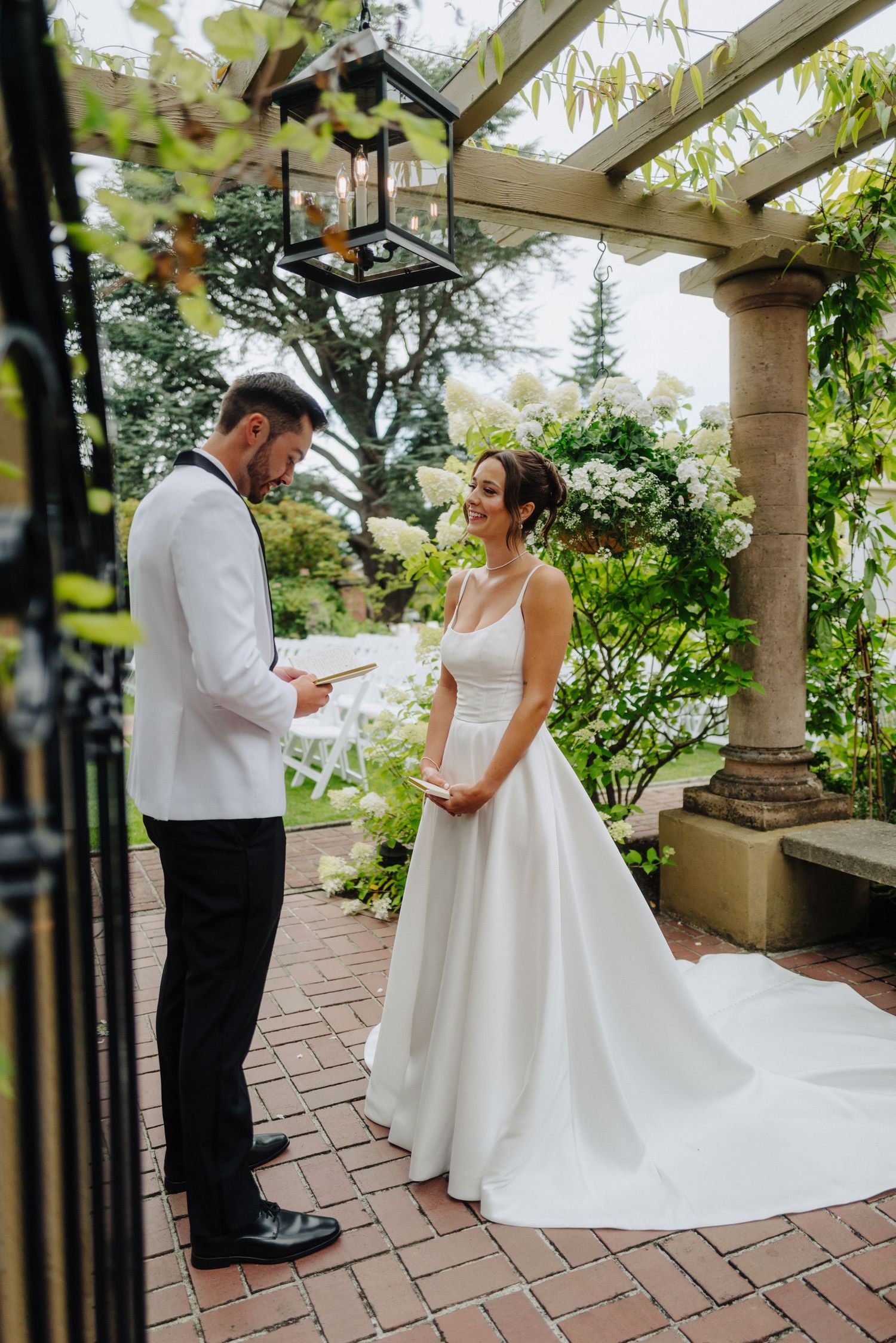 A romantic first look moment captured on a brick patio under a pergola covered in greenery.
