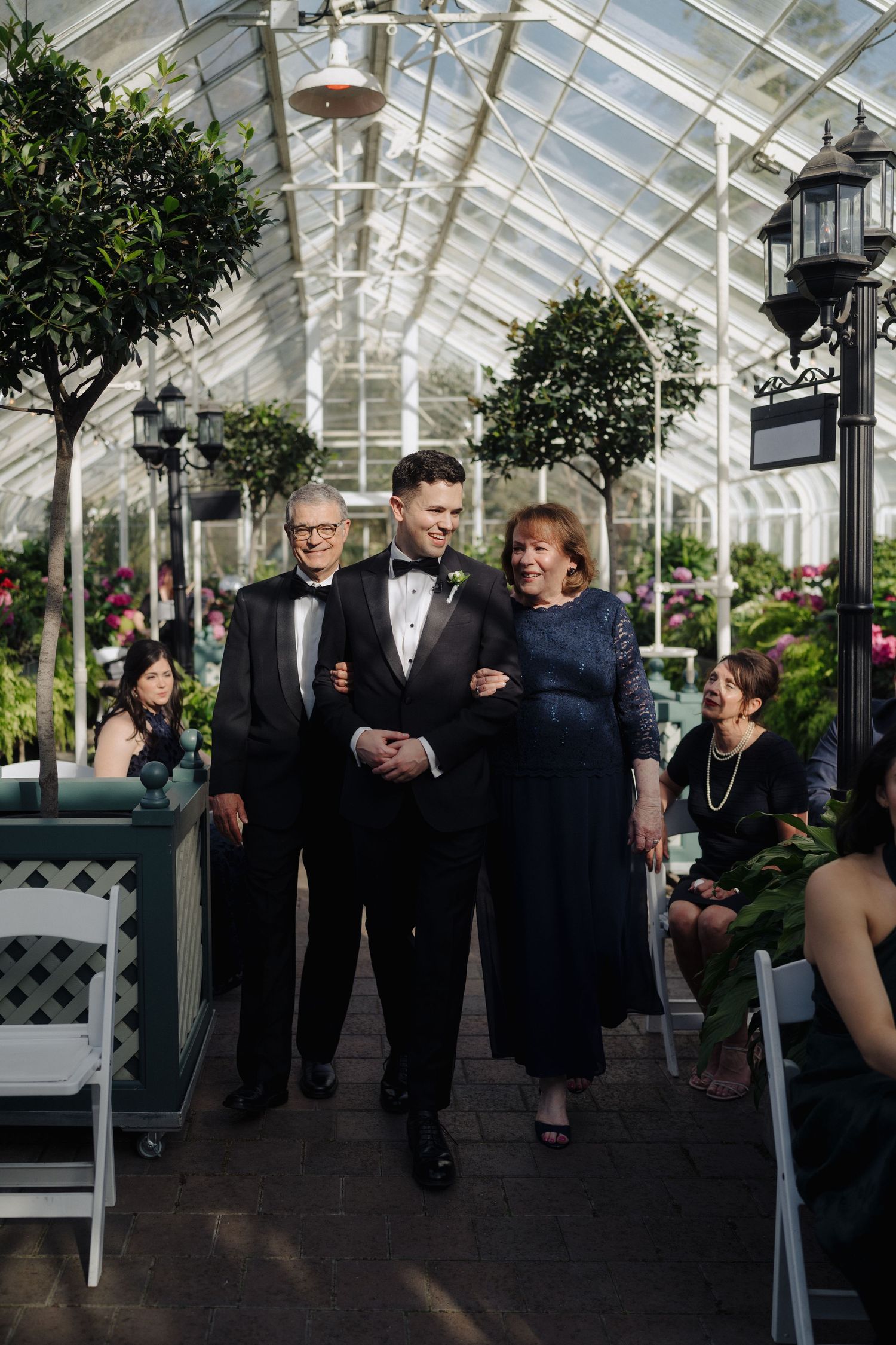 Wedding processional taking place inside a beautiful greenhouse conservatory filled with plants and flowers.