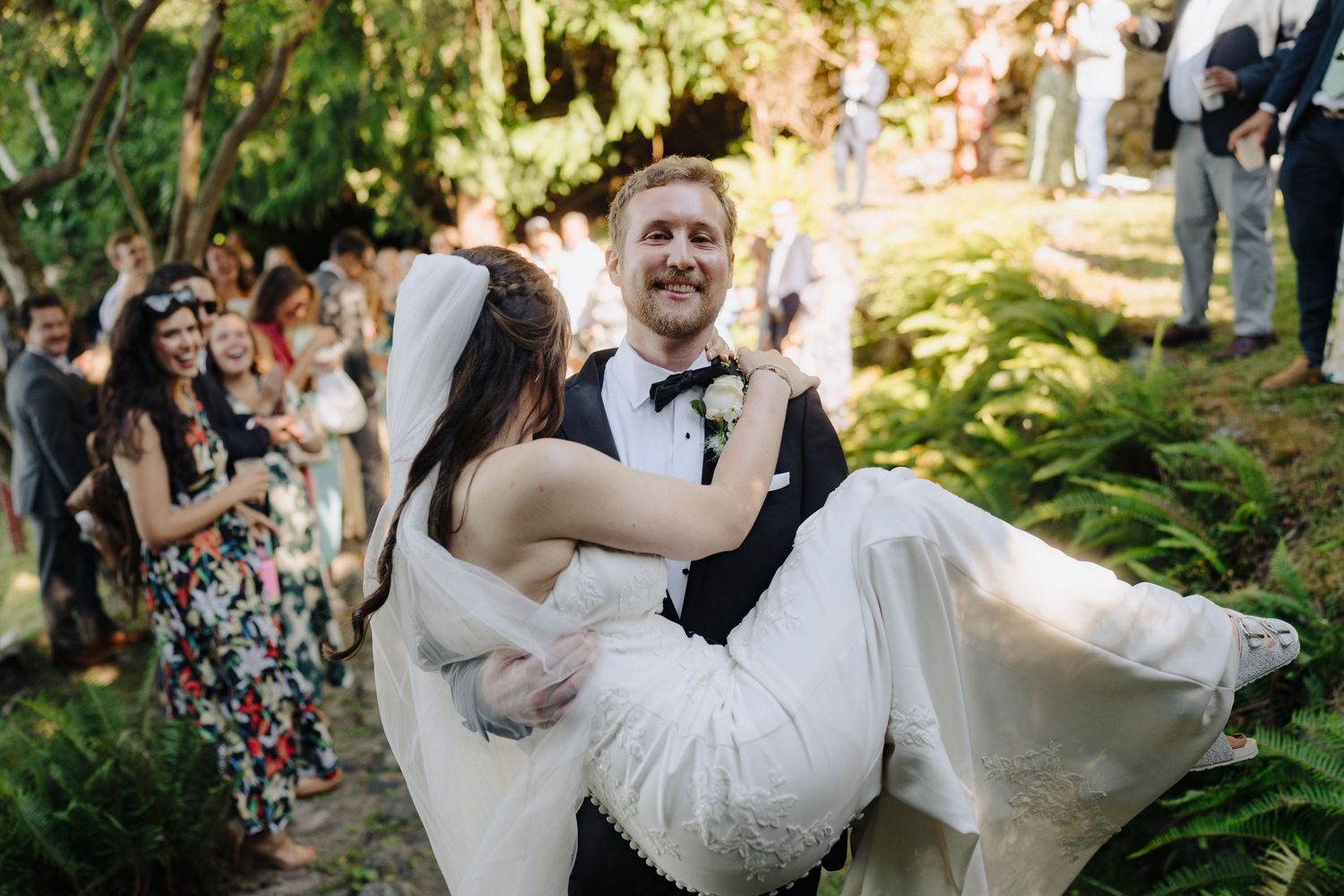 Wedding party celebrates joyfully in an outdoor garden setting with natural greenery and sunlight.