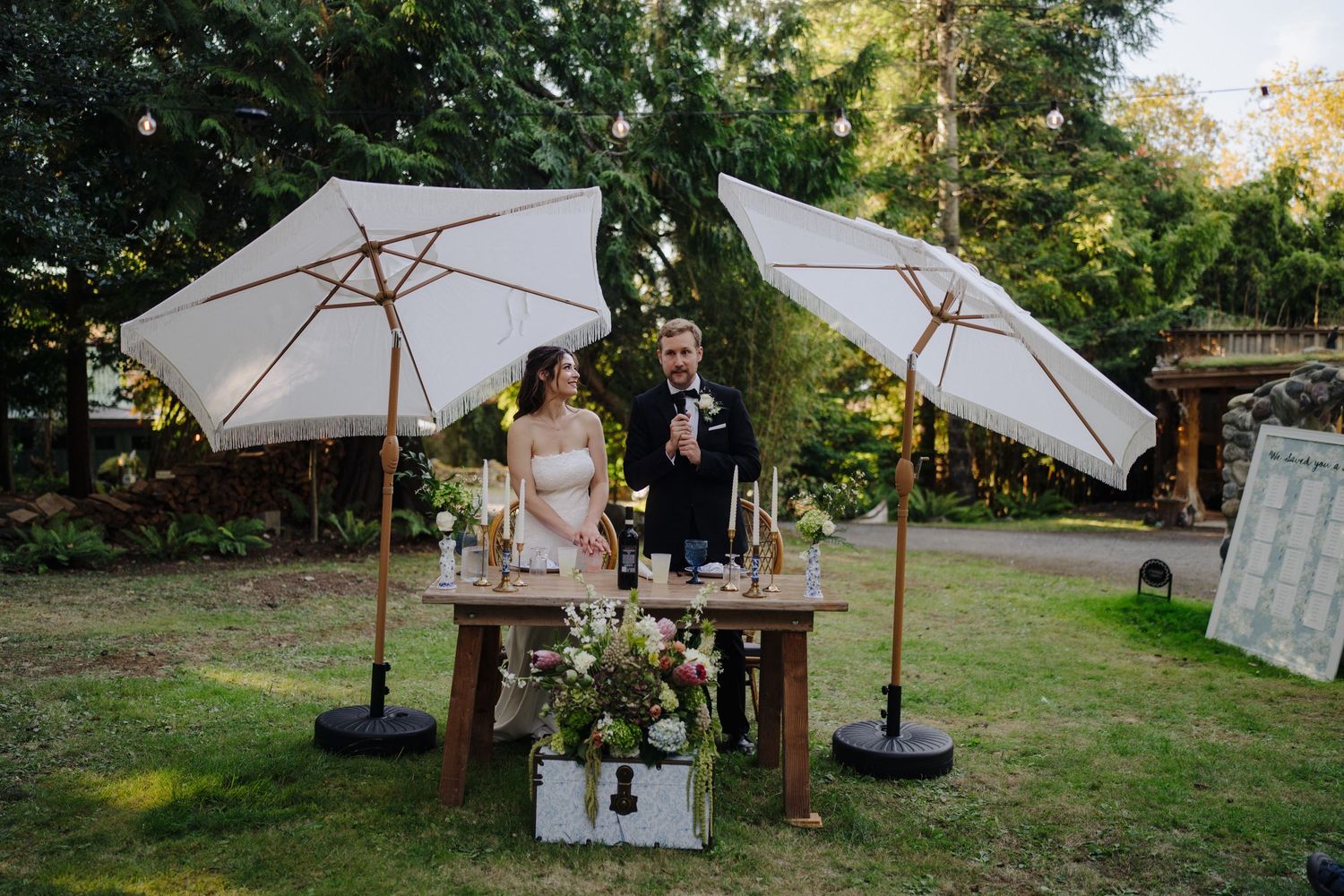 Couple seated at rustic wooden table under white umbrellas during outdoor garden reception.