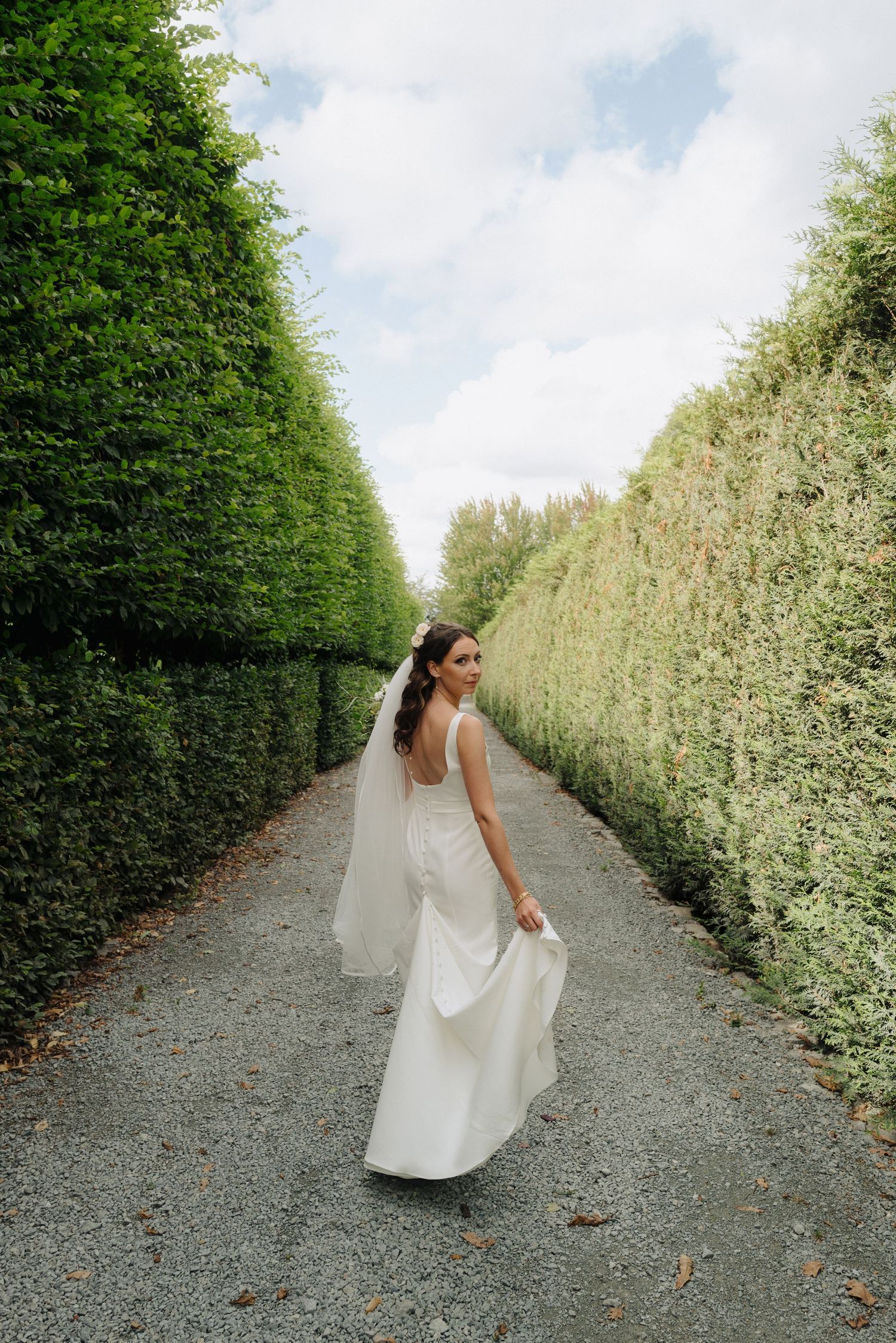 A white dress flows elegantly along a garden path lined with tall green hedges and white flowering bushes.