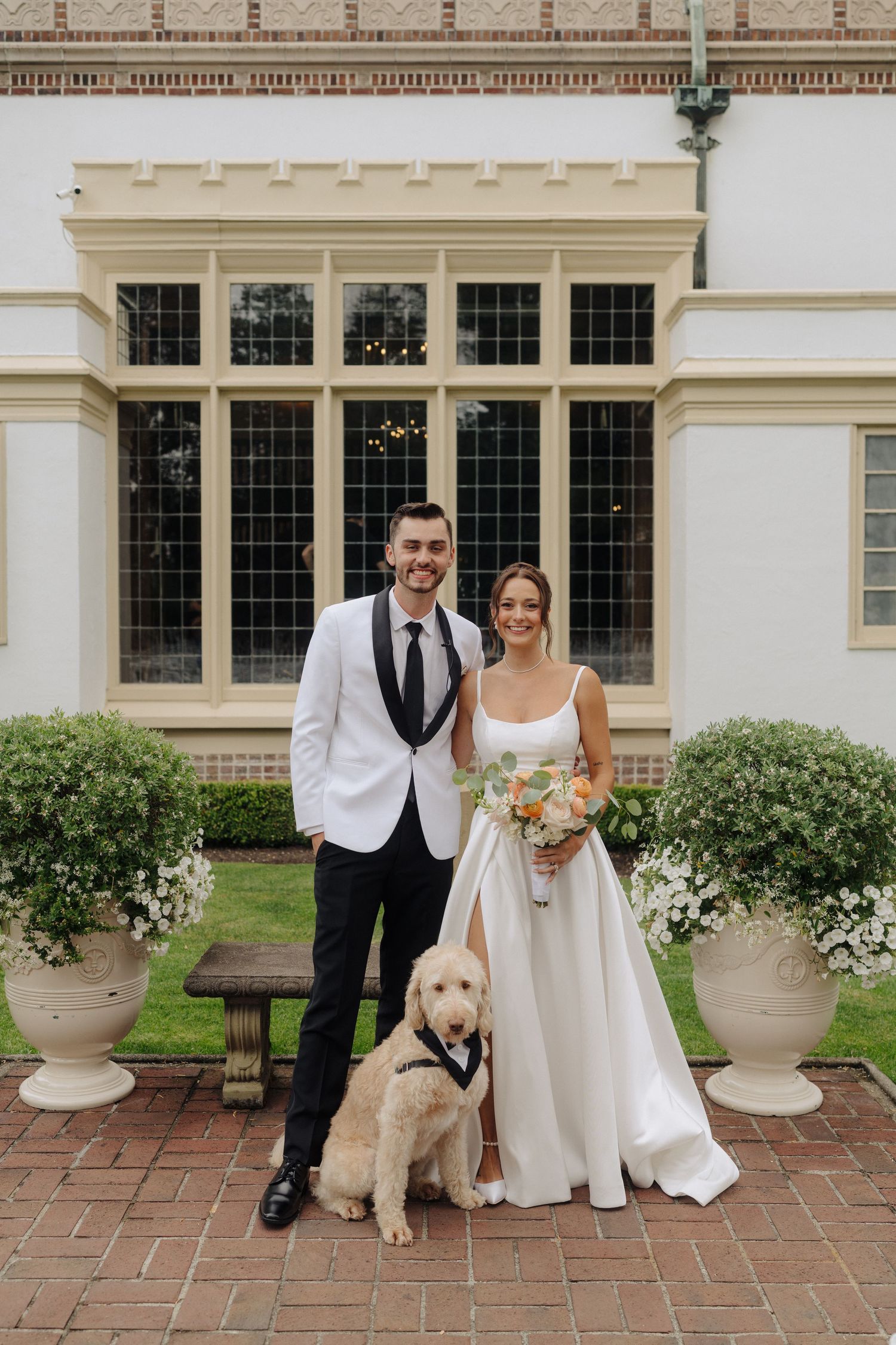 Wedding couple poses with their dog on a brick patio outside an elegant building.