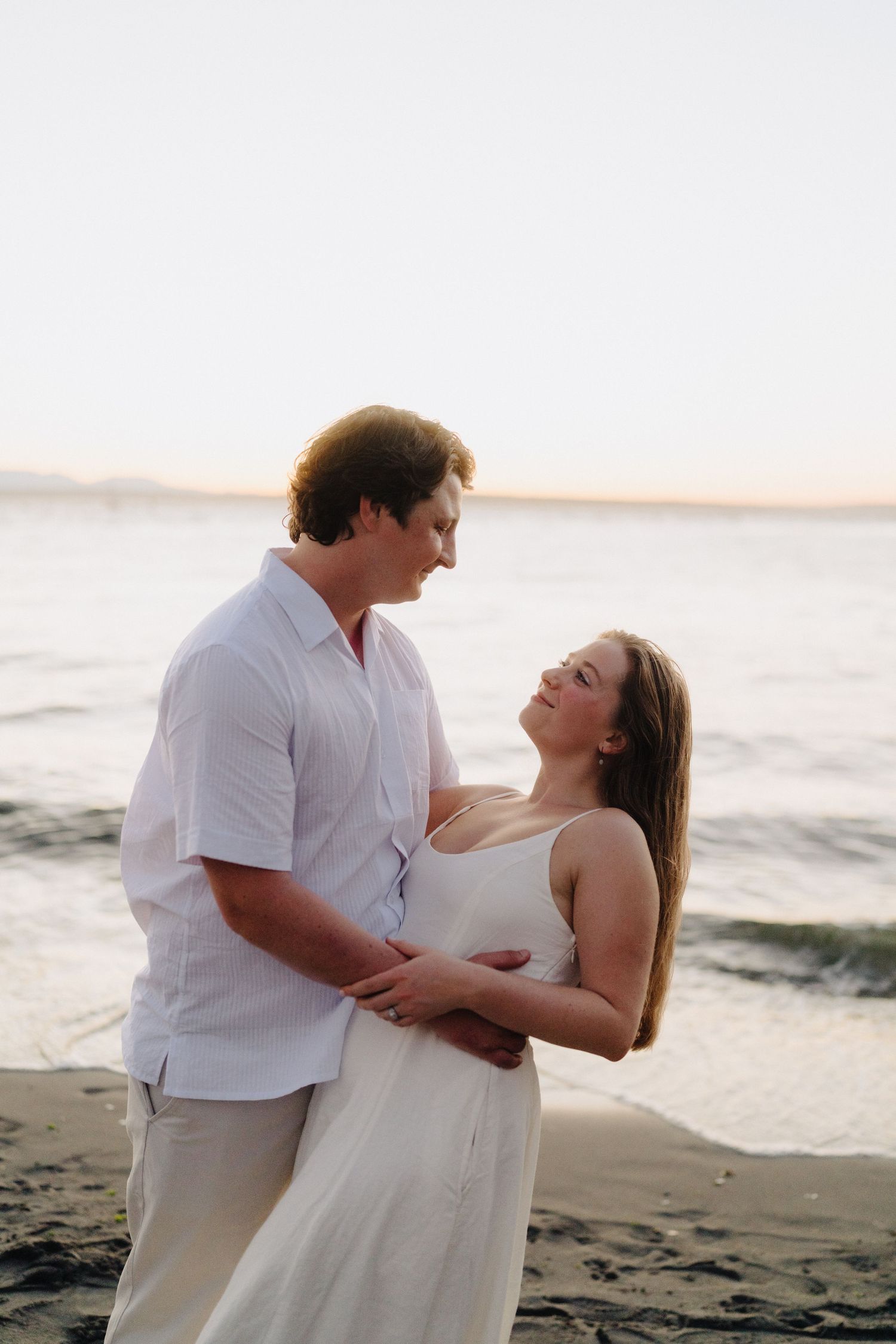 A couple in white clothing embraces romantically on a beach during golden hour.