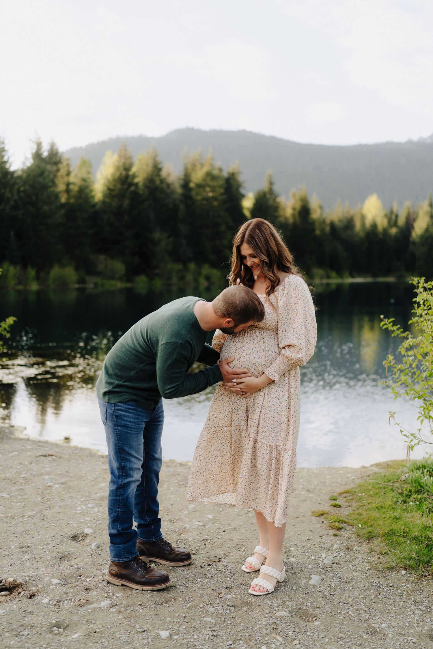 An intimate maternity moment captured by a tranquil lake with mountains and forest in the background.