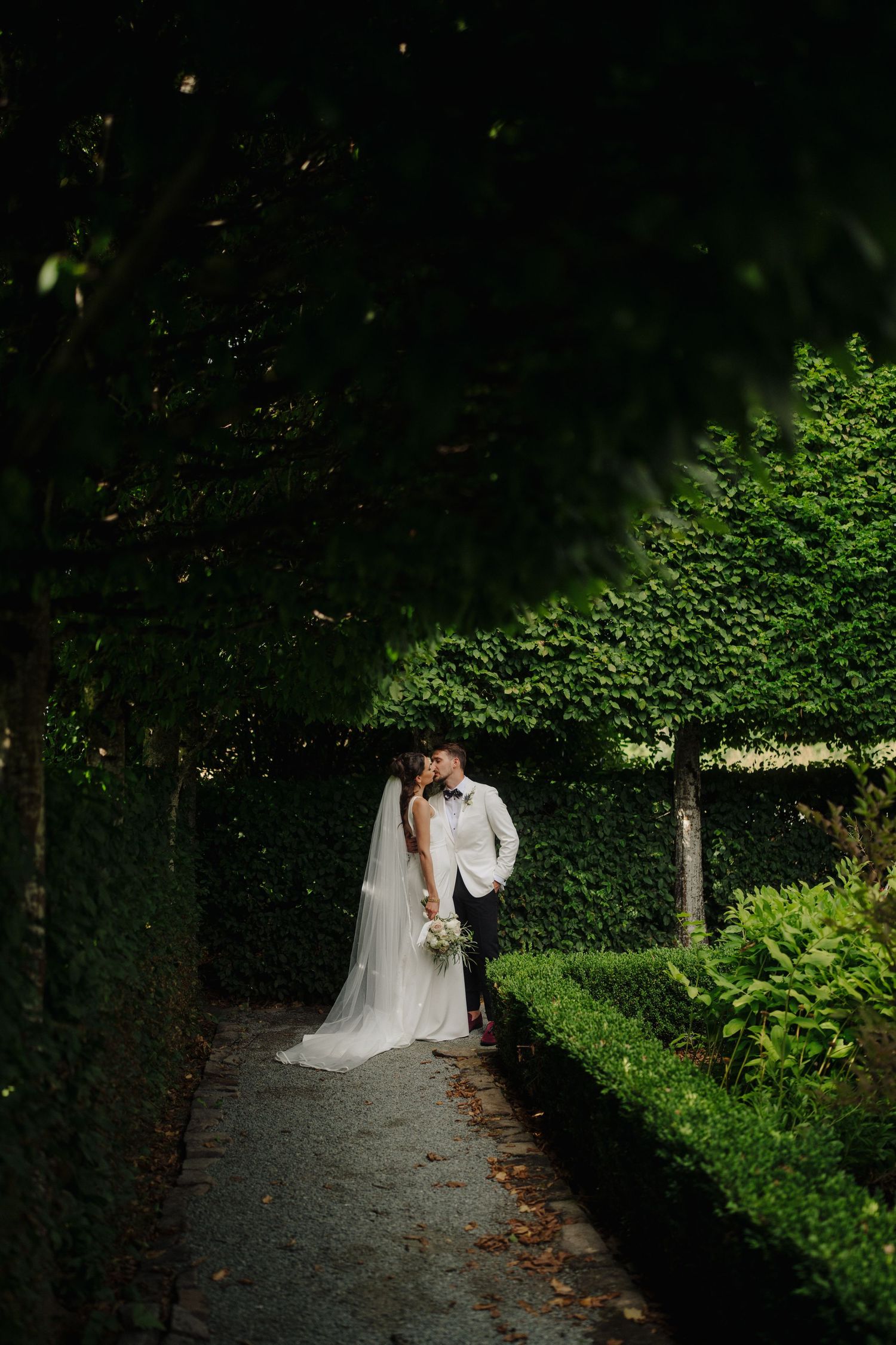 Romantic wedding couple walks along shaded garden path with lush green foliage.