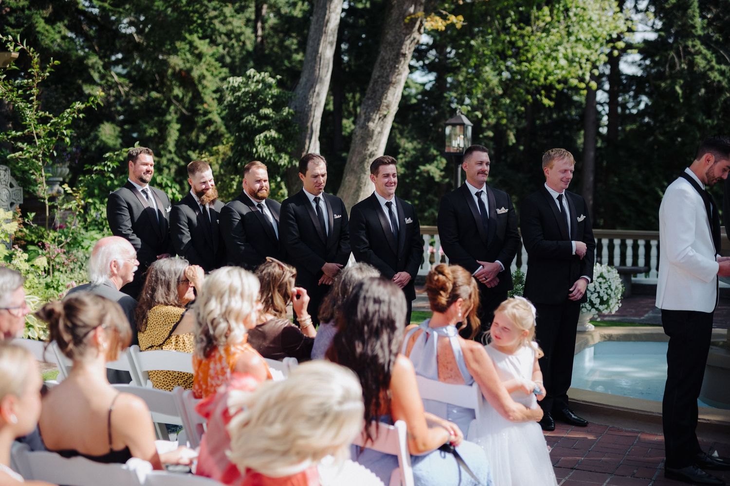 Wedding ceremony guests seated outdoors at a garden venue with groomsmen standing in black suits by trees.