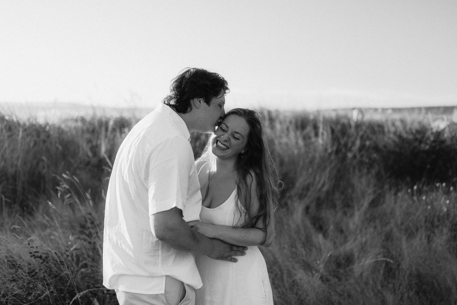 A romantic black and white portrait of a couple embracing in a field of tall grass at sunset.