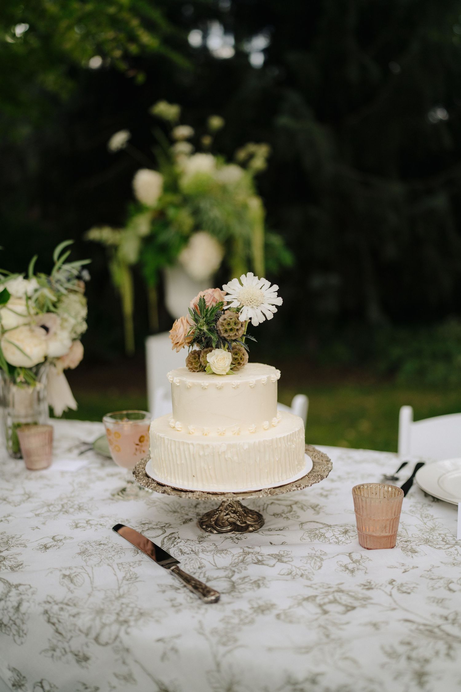 A simple white wedding cake decorated with fresh flowers sits on a patterned tablecloth.
