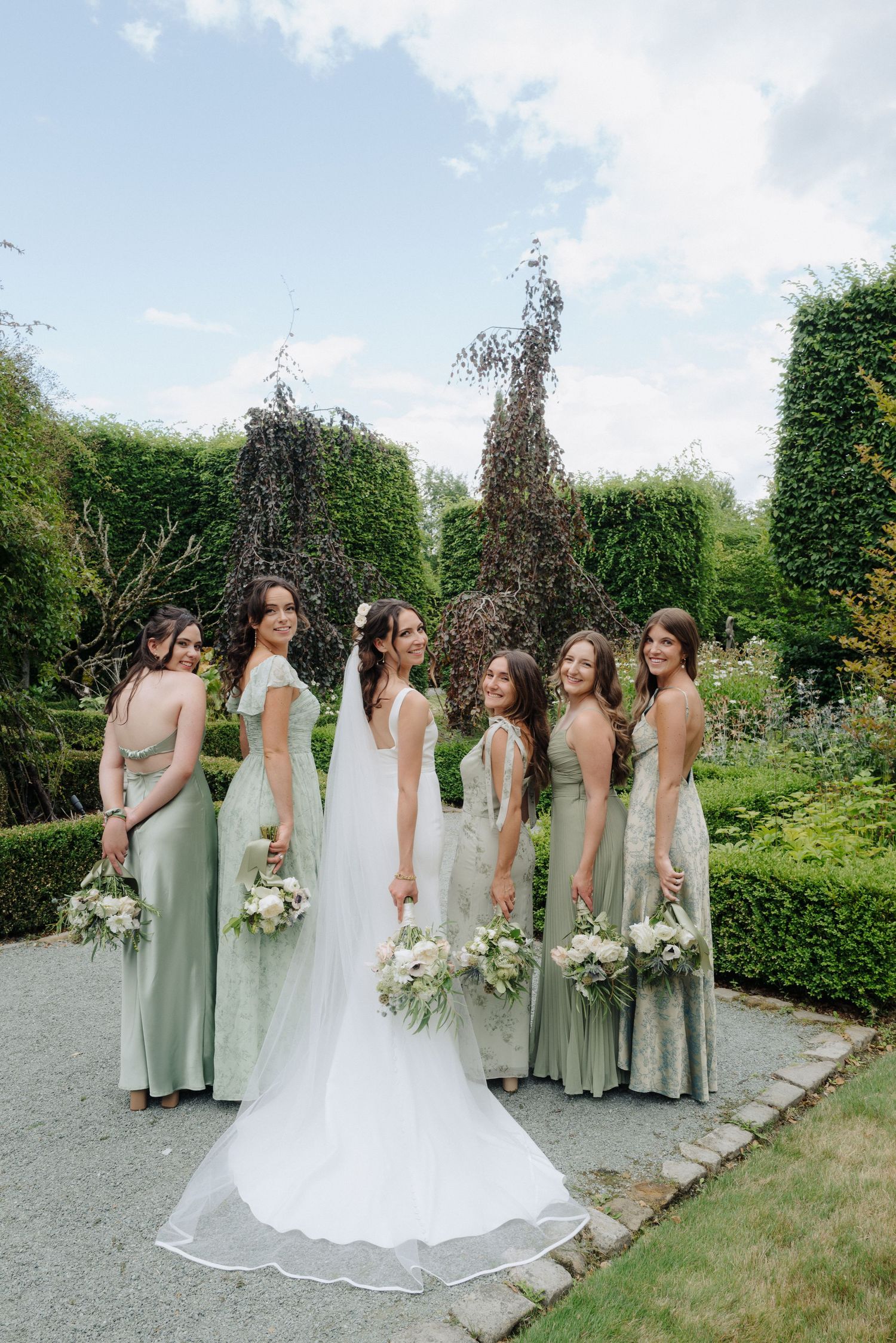 Bridesmaids in sage green dresses pose with bride in flowing white gown against garden backdrop.