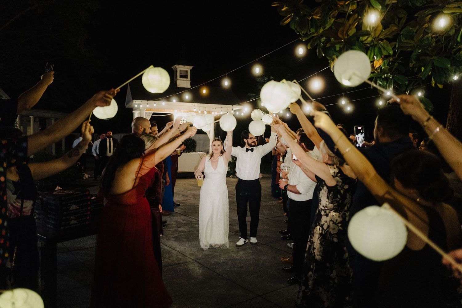Guests celebrate at an outdoor evening reception with glowing paper lanterns and string lights creating a magical ambiance.