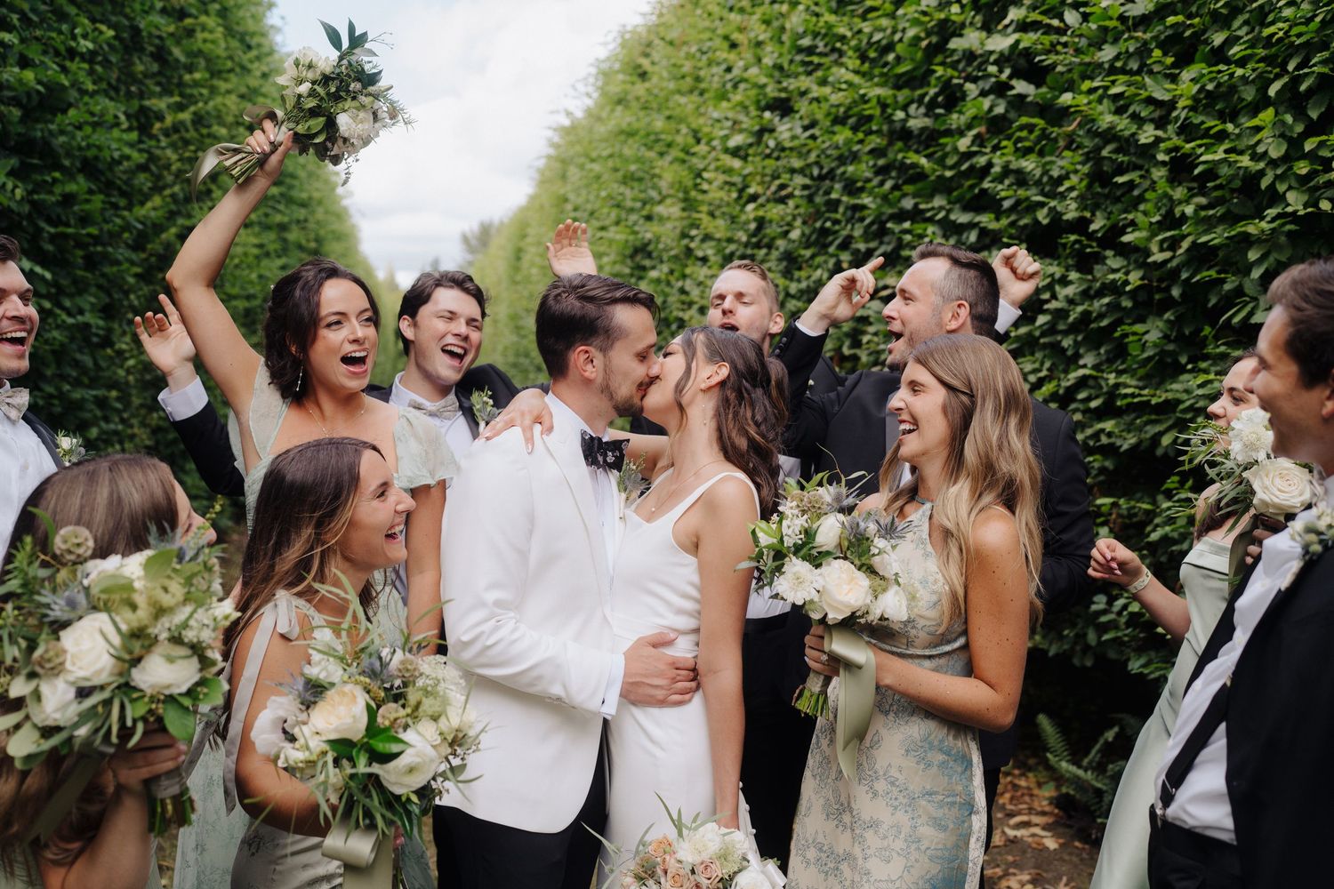Wedding party celebrates joyfully outdoors with raised arms and laughter during summer garden reception.