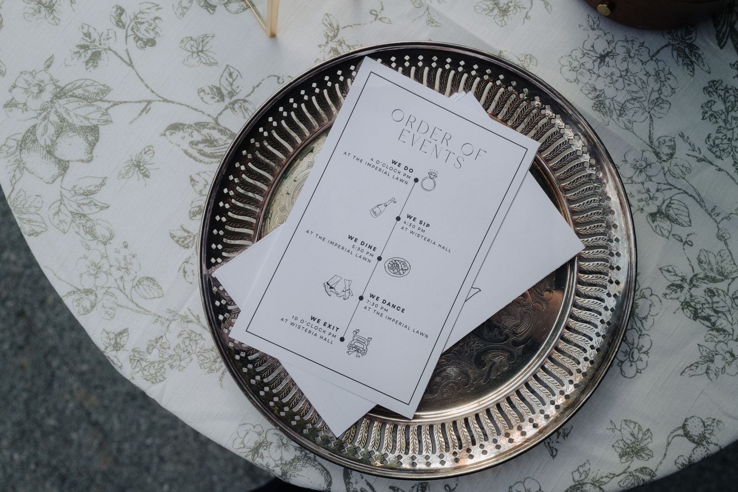 Wedding menu cards displayed on silver decorative plate with floral patterned tablecloth.
