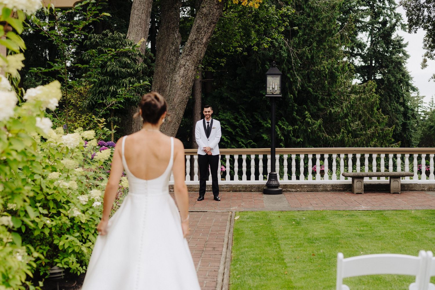 A bride and groom walk together through a lush garden setting with white architectural details in the background.