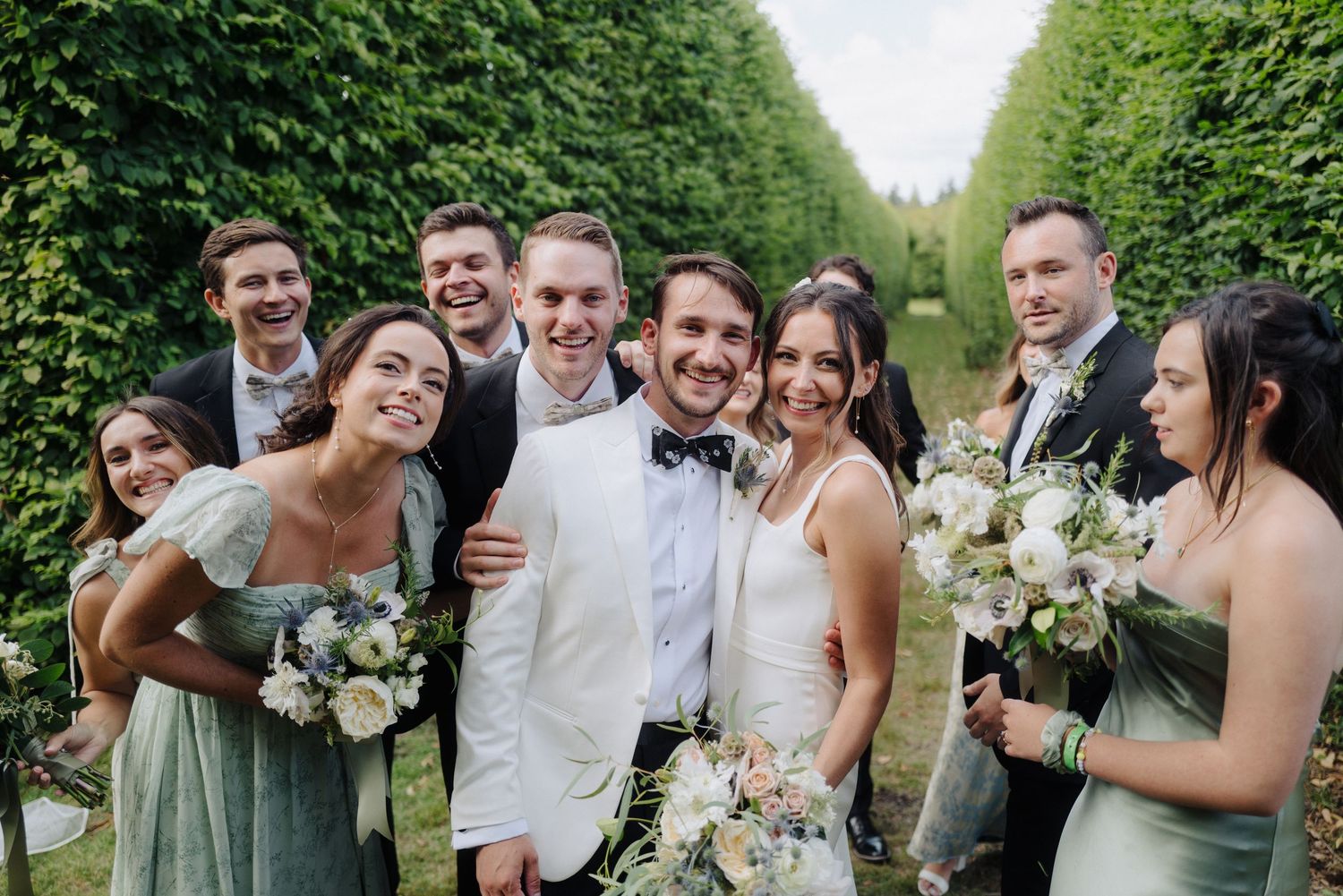 Wedding party members laughing and celebrating in a garden setting with white floral bouquets.