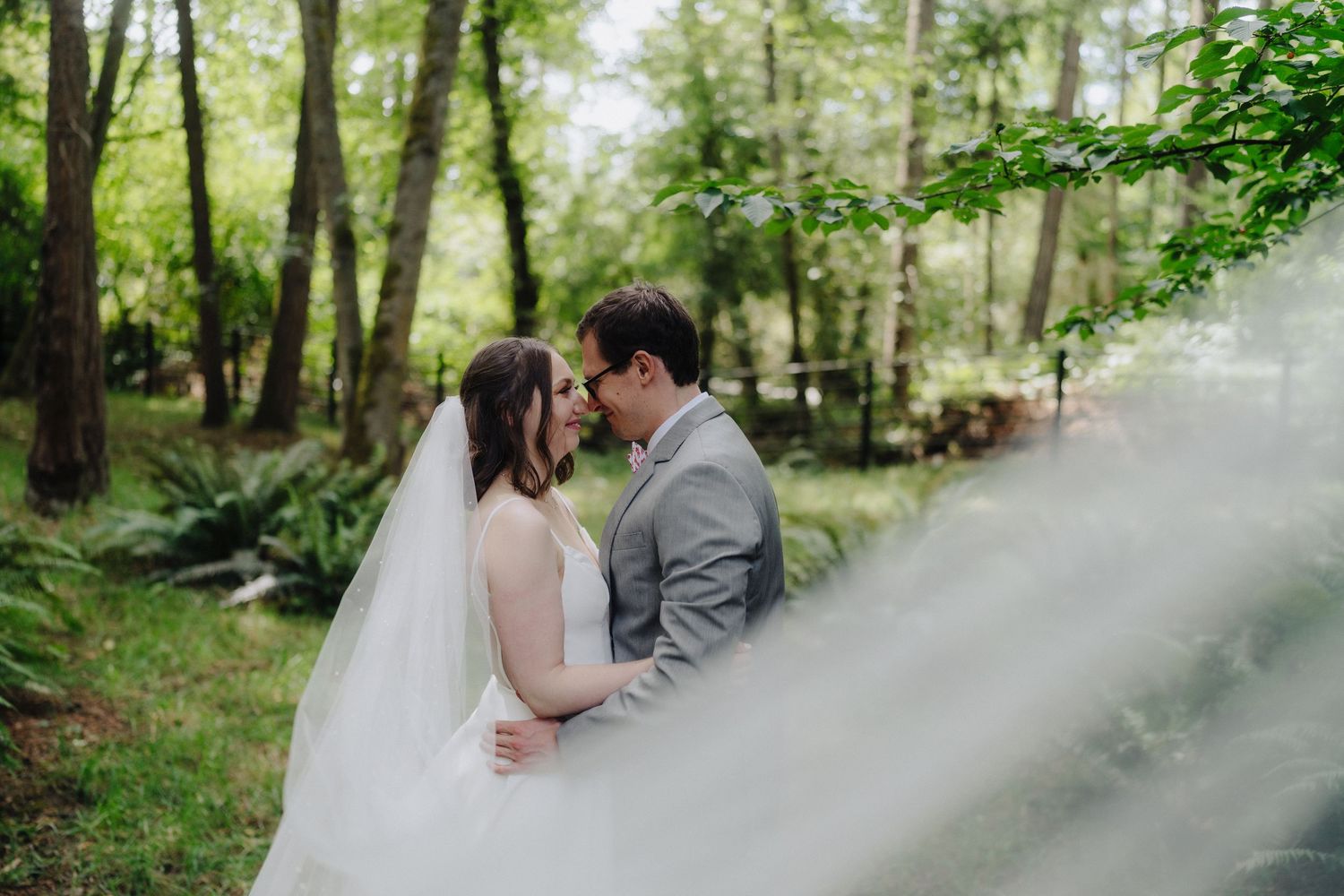 Elegant wedding couple poses in misty forest with flowing white veil.