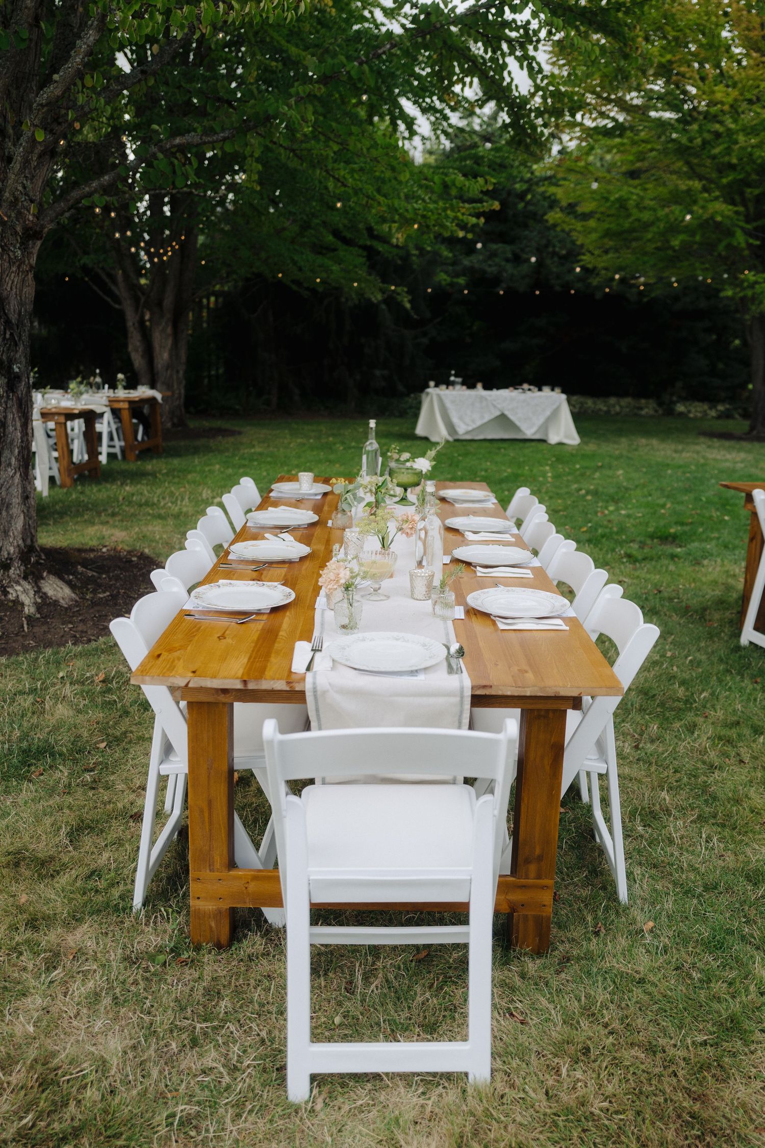 Rustic outdoor wedding reception setup with wooden tables and white chairs on grass.