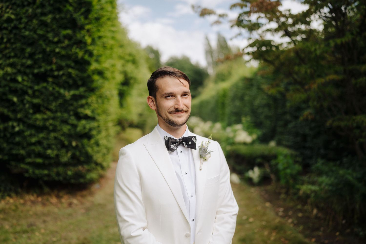 Groom in white tuxedo and bowtie poses in garden pathway.