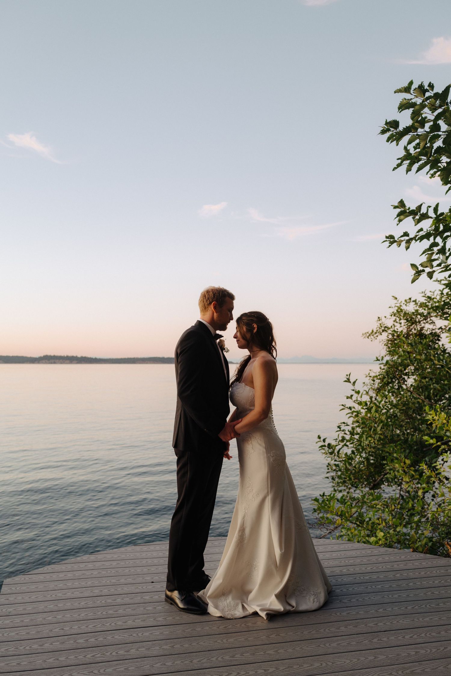 A couple in wedding attire shares an intimate moment on a wooden deck overlooking a serene lake at sunset.
