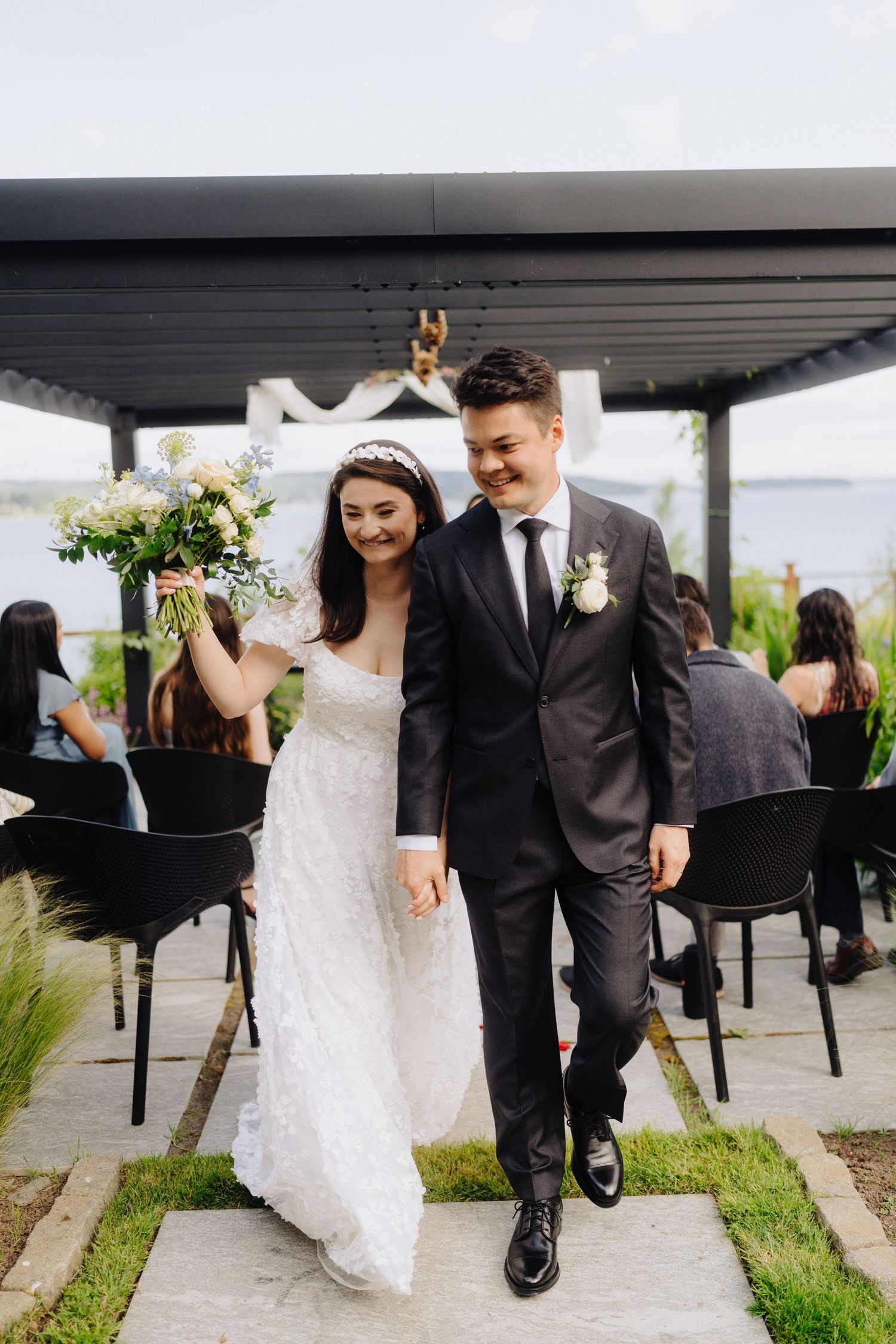 A joyful wedding couple walks together under a covered outdoor deck by the water.