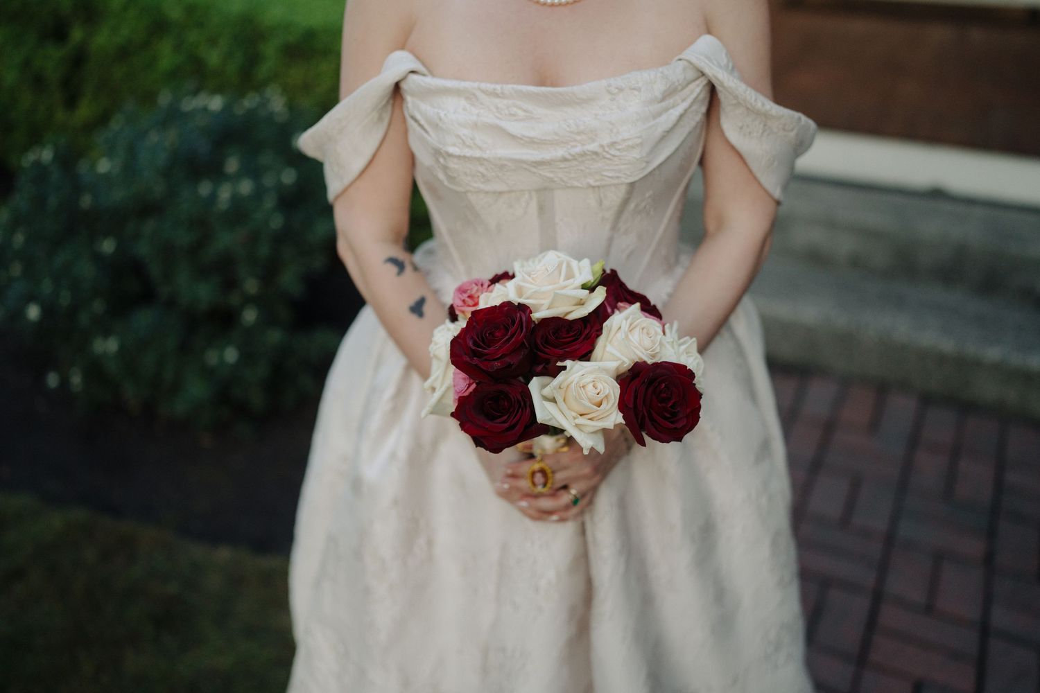 White wedding dress with red and burgundy bouquet details against a brick pathway backdrop.