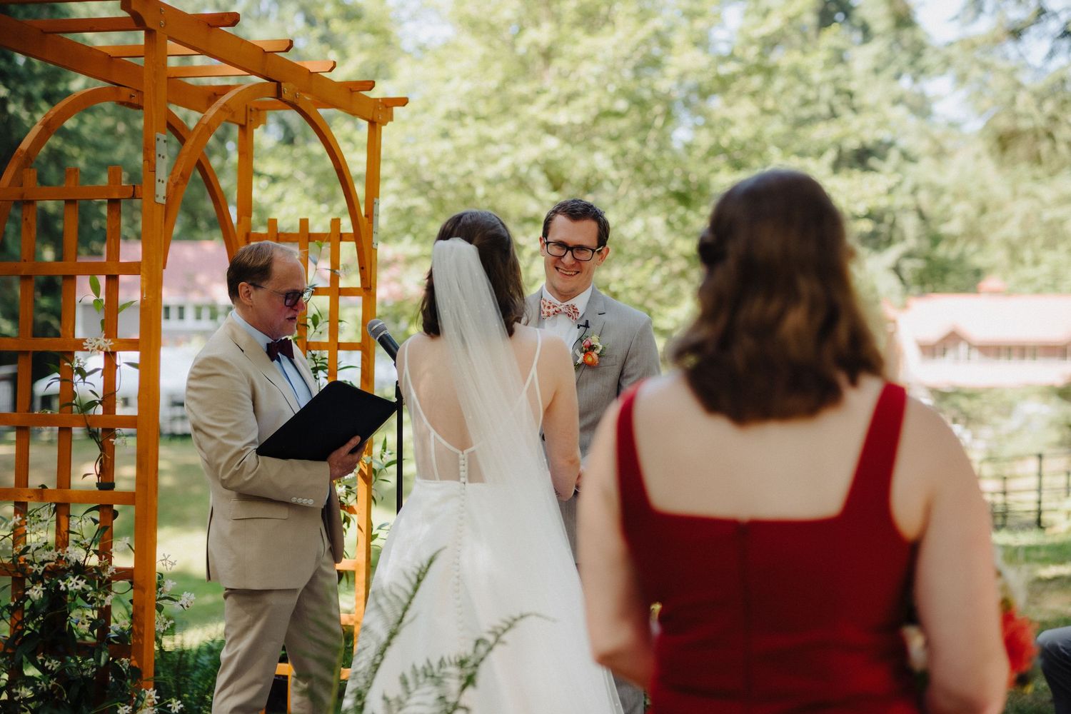 Wedding ceremony taking place under a wooden pergola surrounded by trees and guests in an outdoor garden setting.