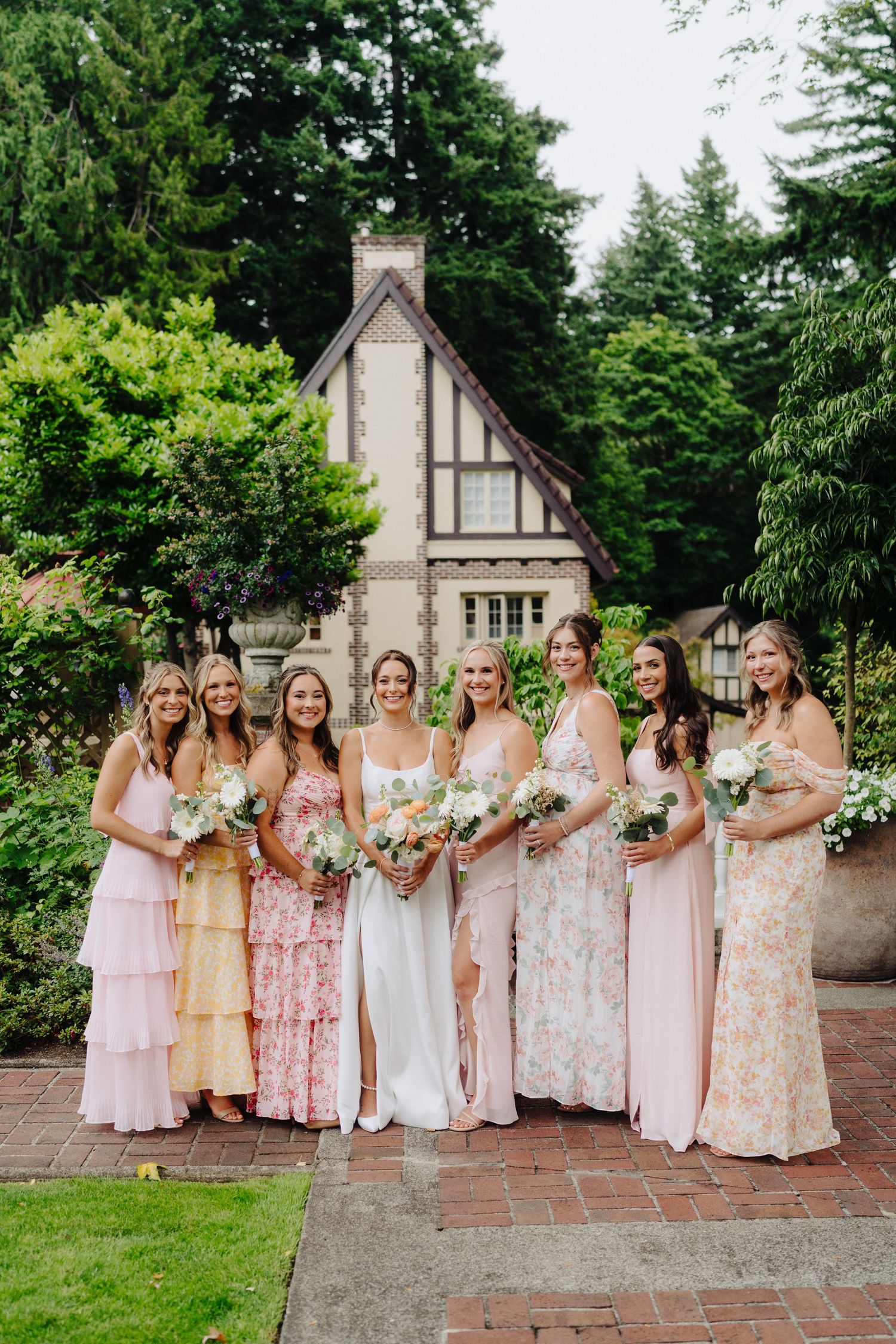 A group of bridesmaids in soft pink and floral dresses pose together on brick steps at a garden wedding venue.