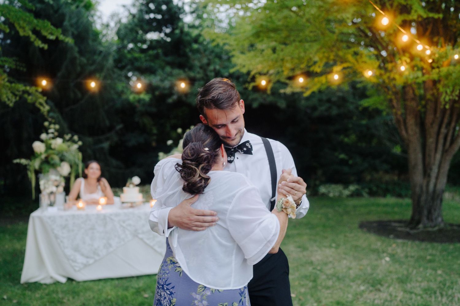 Couple dancing at twilight outdoor reception with string lights overhead.