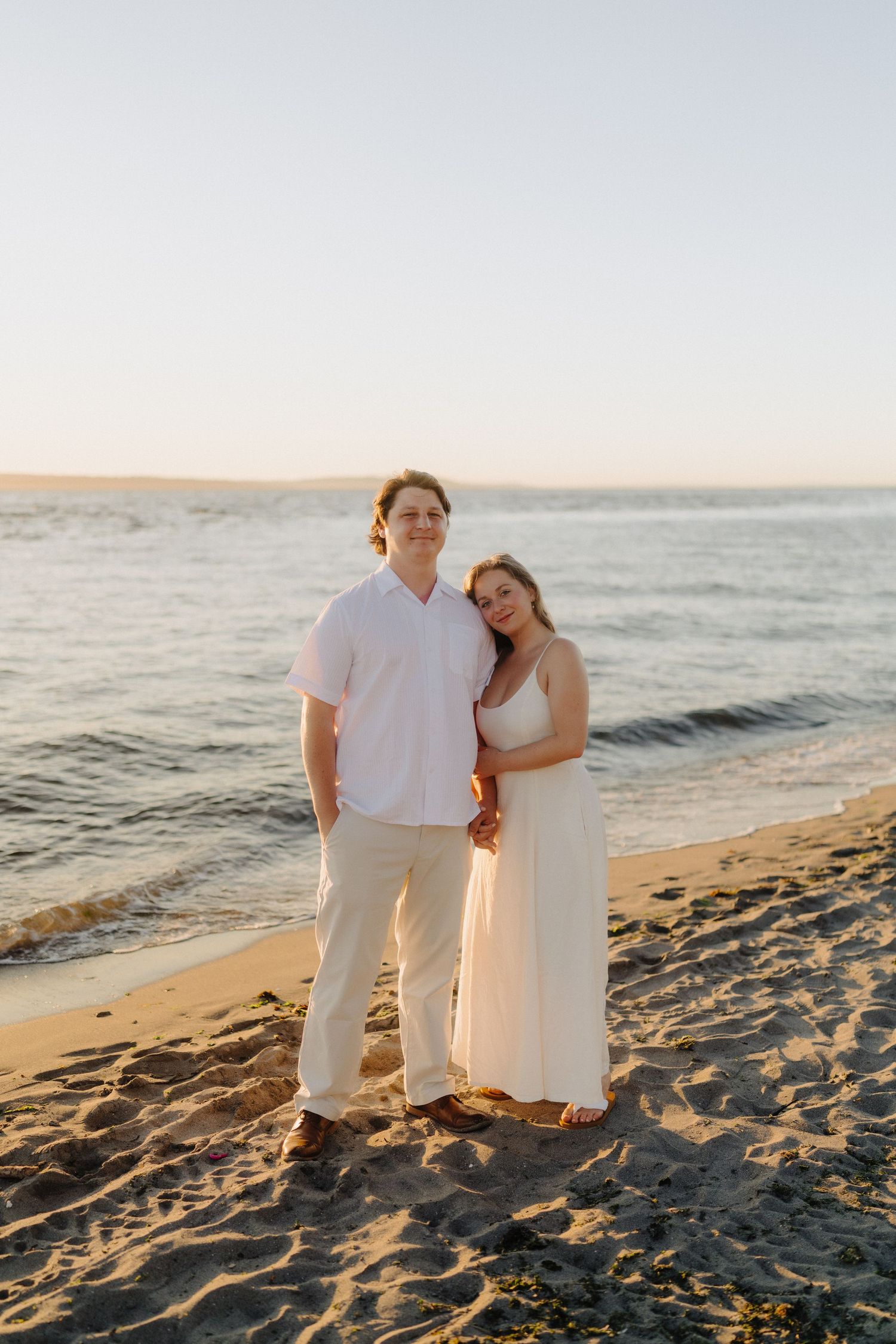 A couple dressed in white embraces on a sandy beach at sunset with ocean waves in the background.