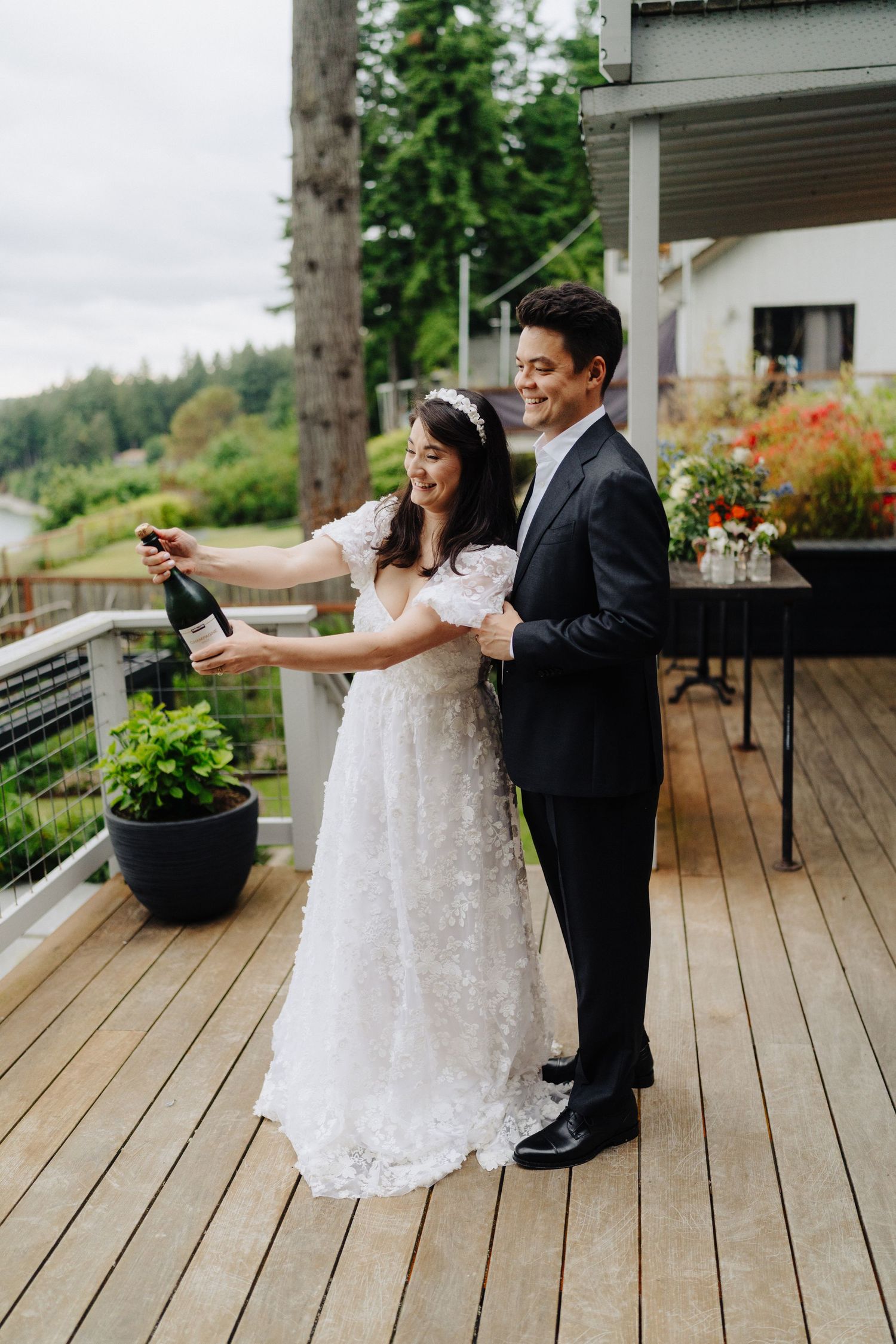 A couple celebrates with champagne on a wooden deck overlooking a forested hillside.