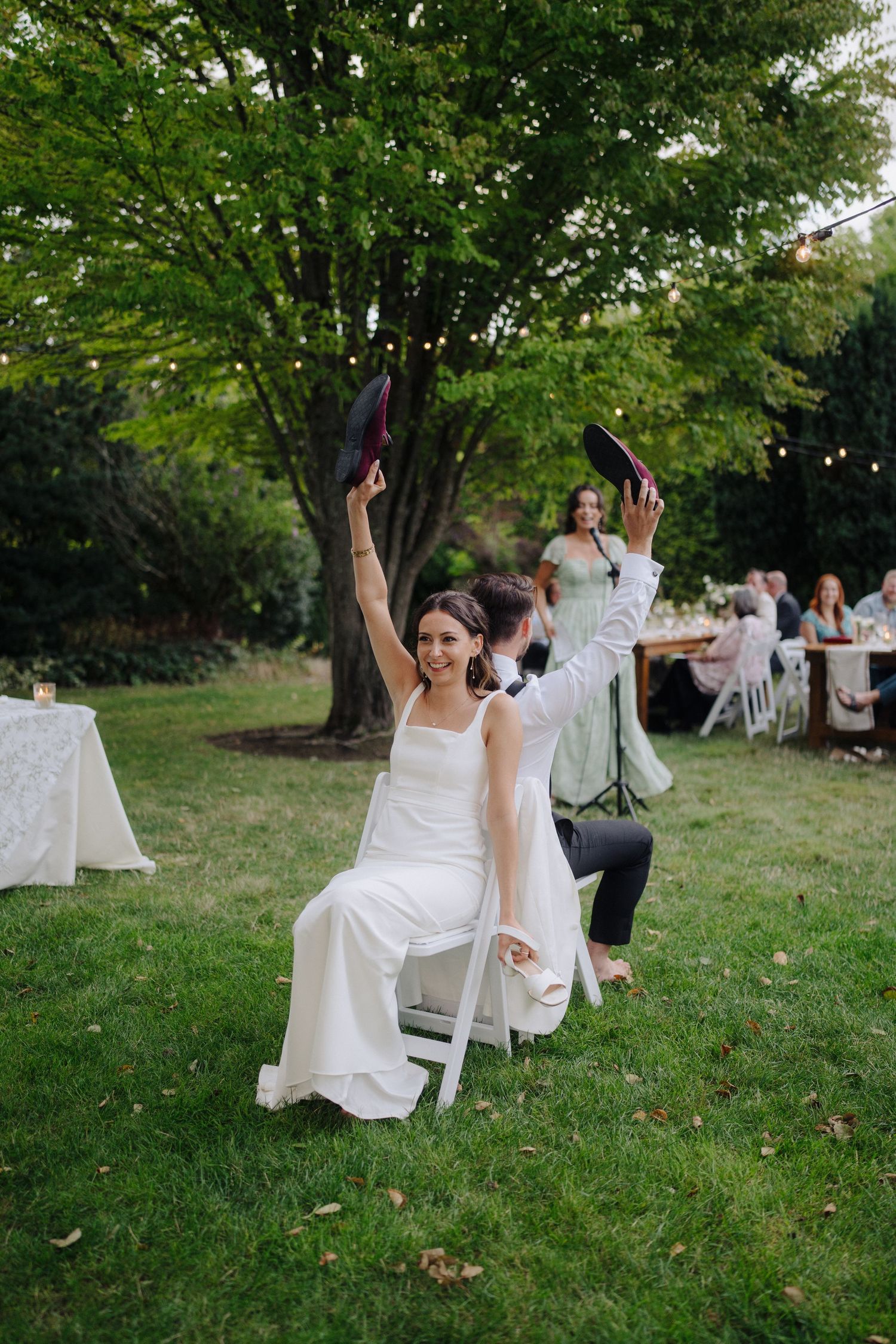 Wedding guests enjoy outdoor reception seating on white chairs under trees.