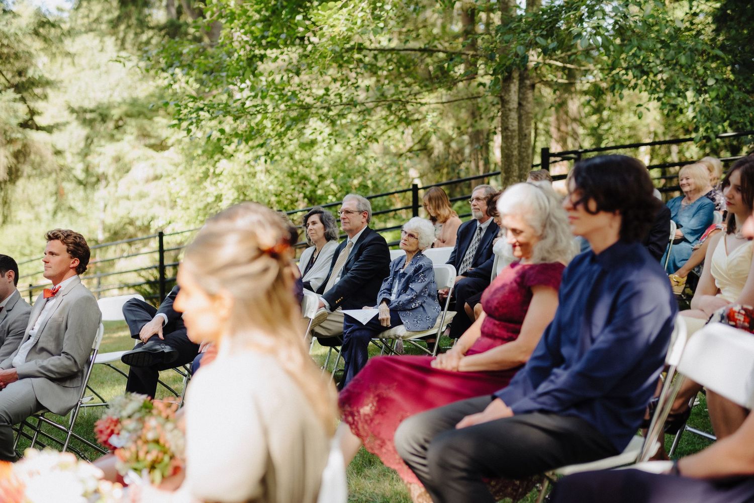 Wedding guests seated in white chairs watching outdoor ceremony in a wooded setting.
