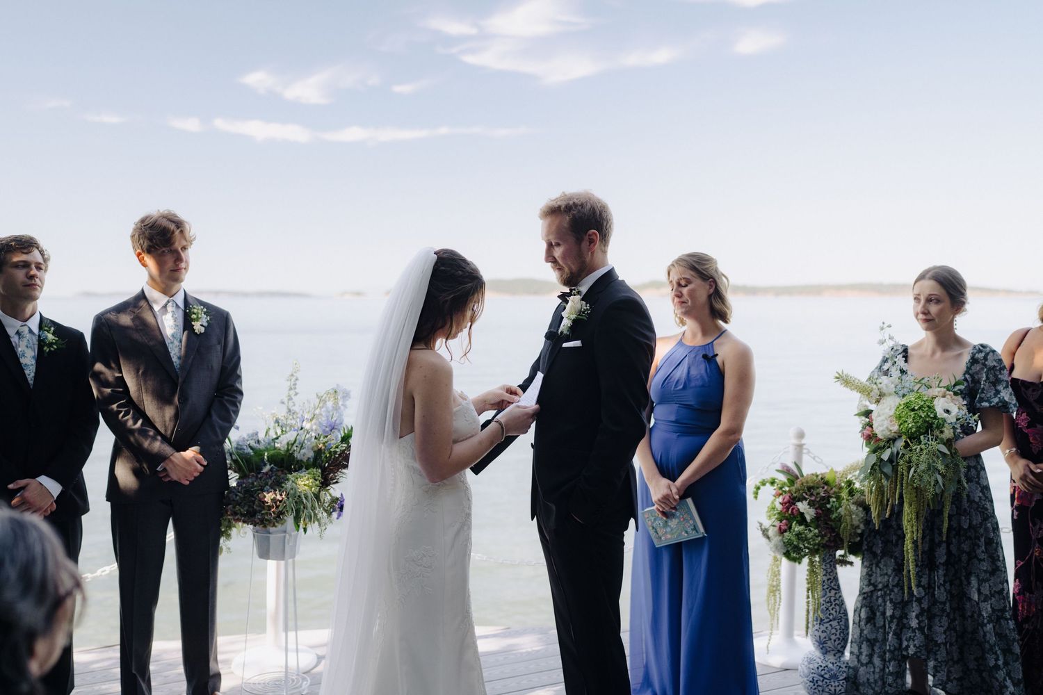 Wedding ceremony participants stand in line during outdoor beach ceremony with ocean view in background.